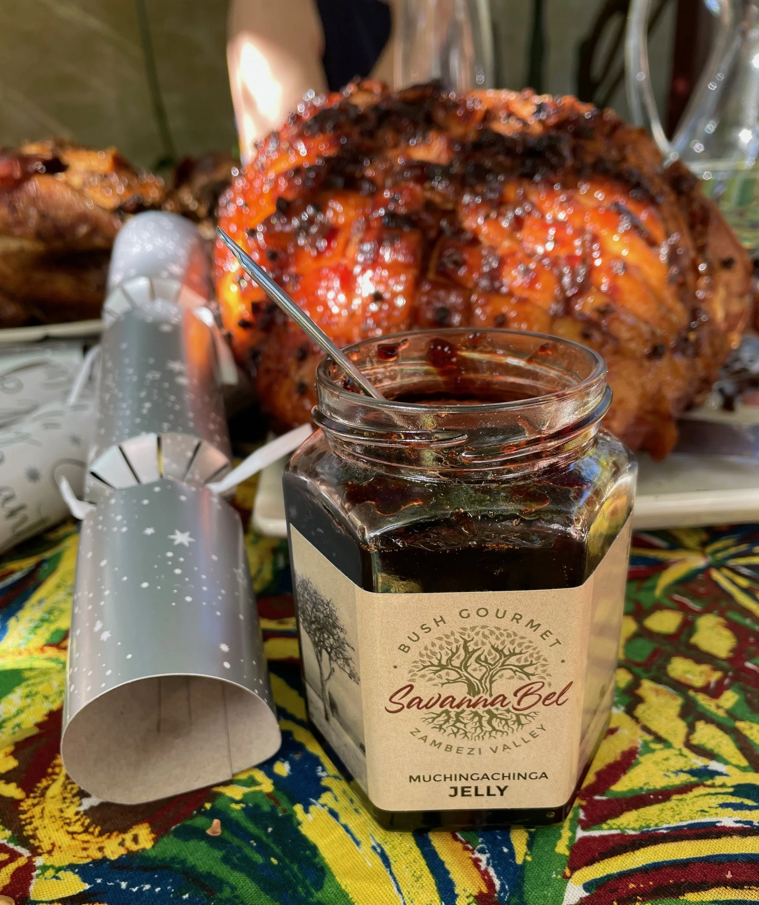 A jar of Bush Gourmet Savannah Bell Muchingachinga Jelly in front of a large roasted dish with vegetables, on a colorful tablecloth.