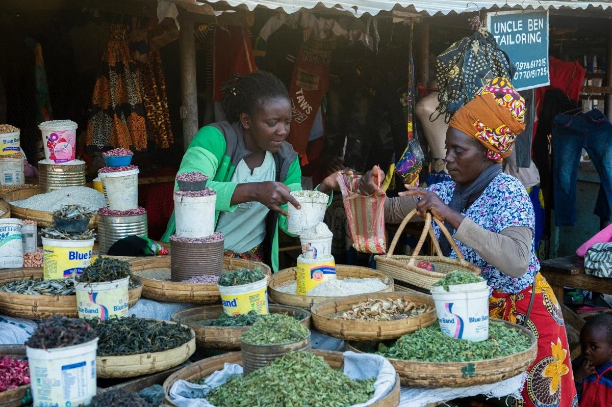 Two women engaged in a transaction at a market stall filled with various spices and herbs displayed in round woven baskets. The woman on the left, wearing a green and gray jacket, is serving a customer, the woman on the right, who is wearing an orange headwrap and a colorful dress. Behind them, there is a sign that reads 'Uncle Ben Tailoring,' indicating nearby tailoring services.