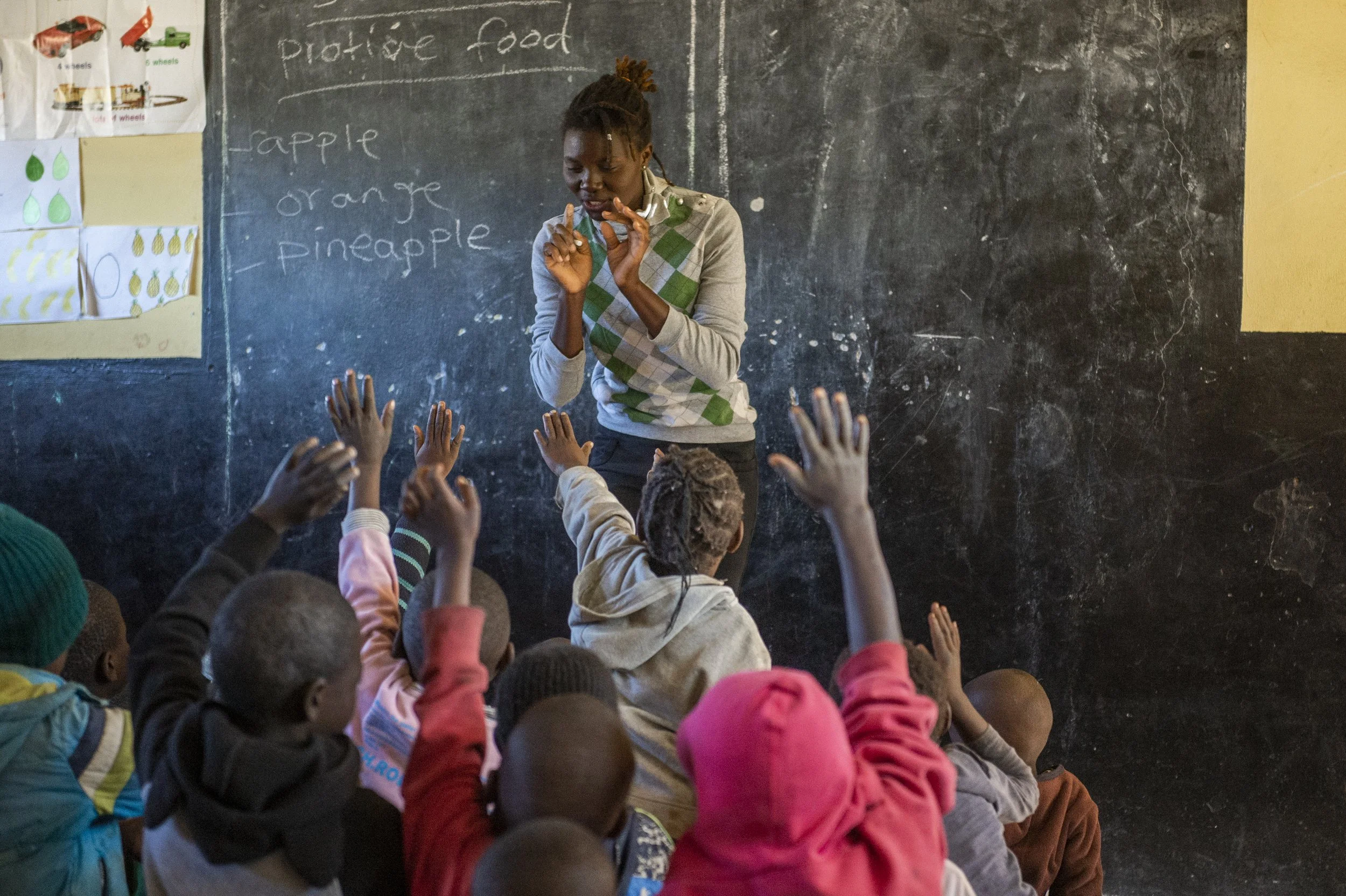 A classroom scene with a teacher standing in front of a blackboard and raising her hands in a teaching gesture, while children sit on the floor with their hands raised, listening attentively. The blackboard has written words related to fruit.
