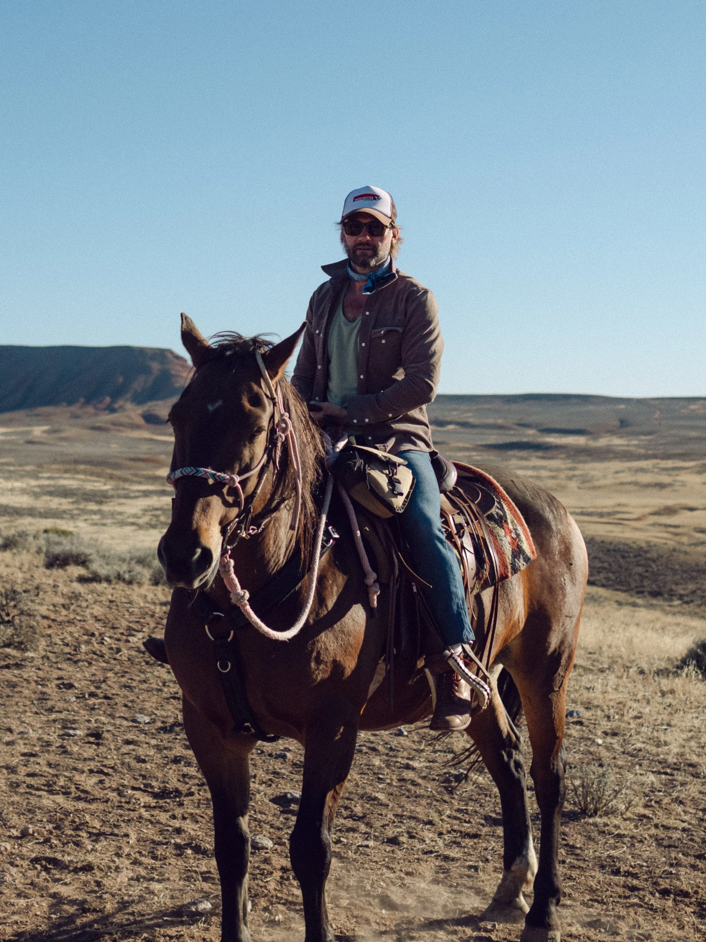 A man wearing a brown jacket, sunglasses, and a cap riding a brown horse across a dry, open landscape with hills in the background under a clear blue sky.