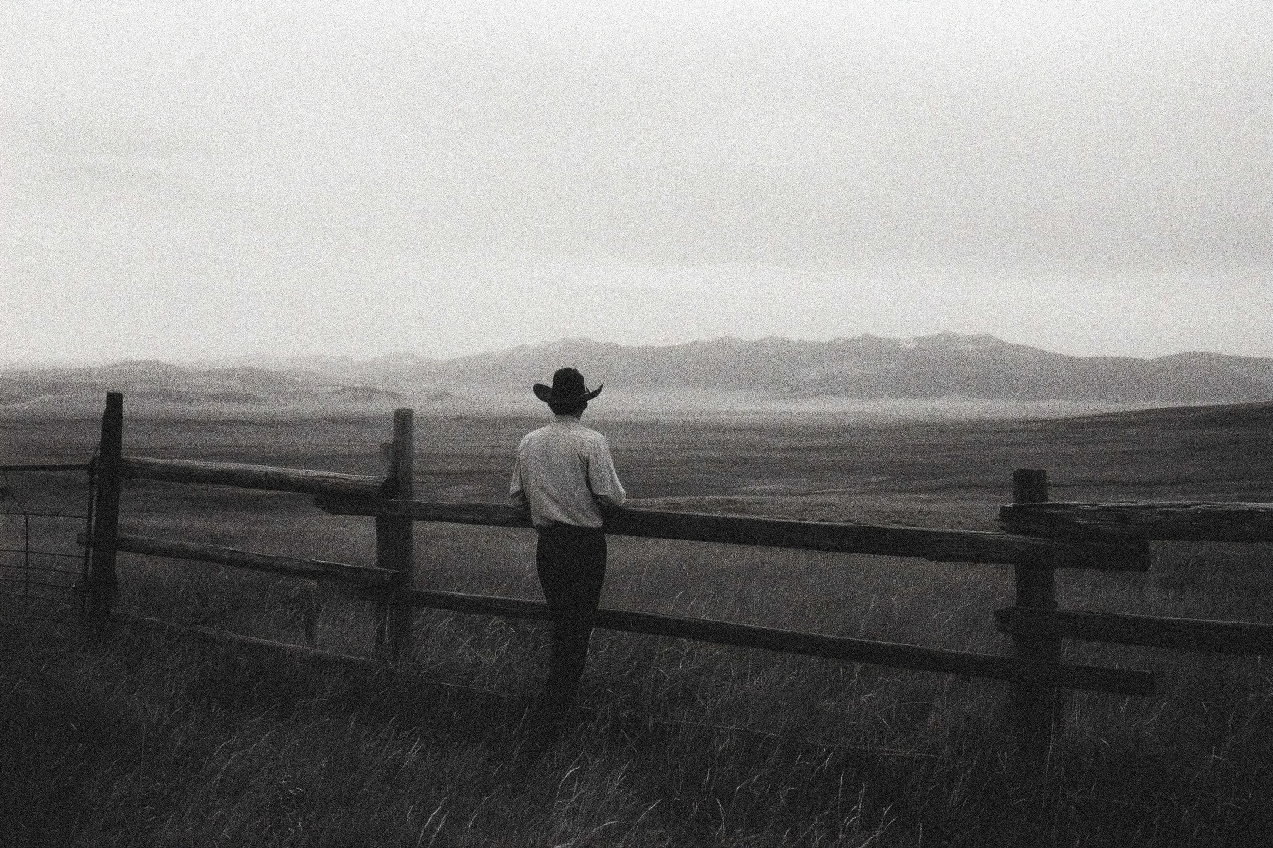 A person wearing a cowboy hat and white shirt standing by a fence, looking out over a landscape with mountains in the distance in black and white.