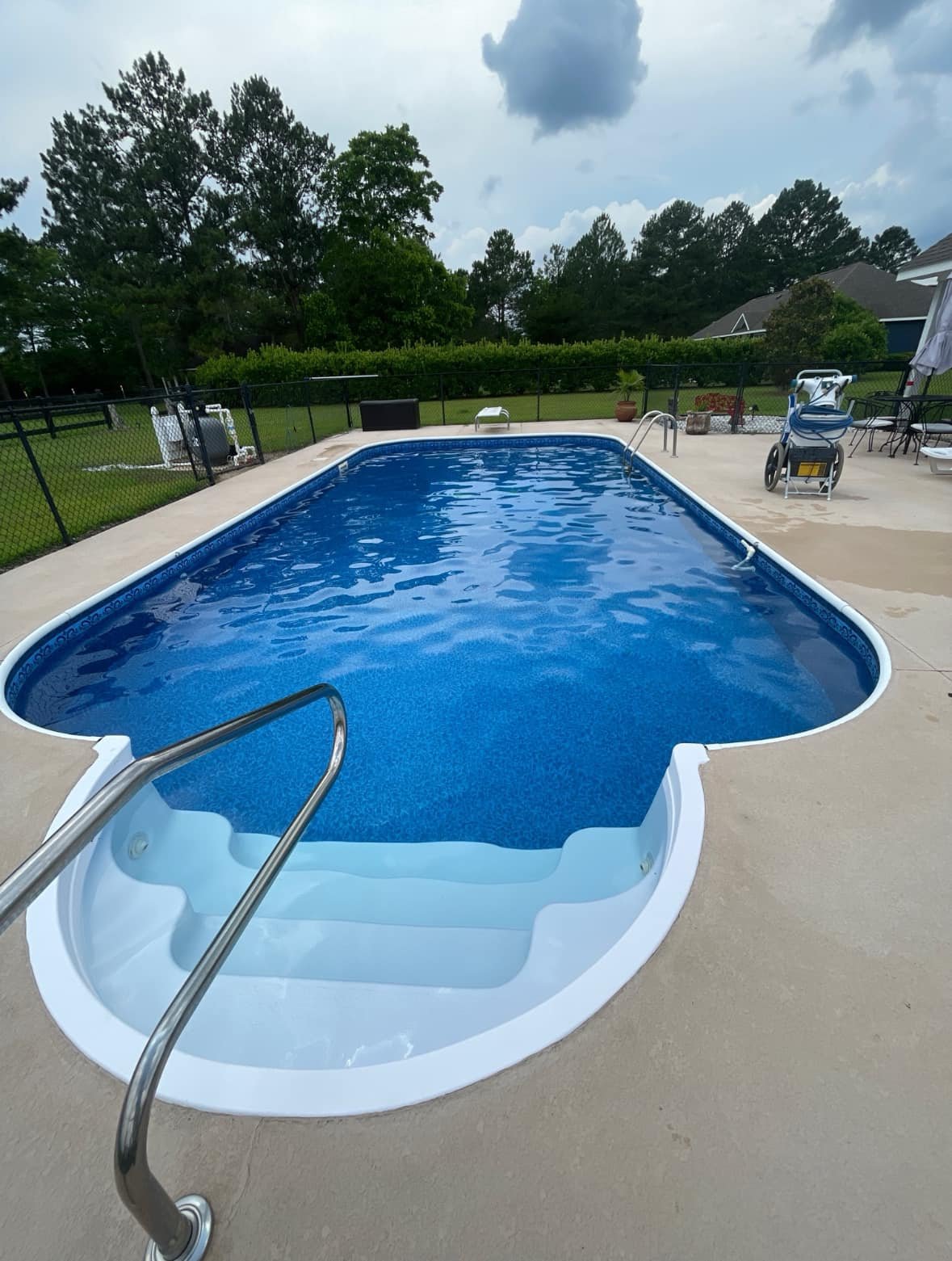 A backyard swimming pool with a white frame and blue water, surrounded by a concrete deck. There are steps leading into the pool, and a metal handrail. The background features a black fence, trees, and outdoor furniture.