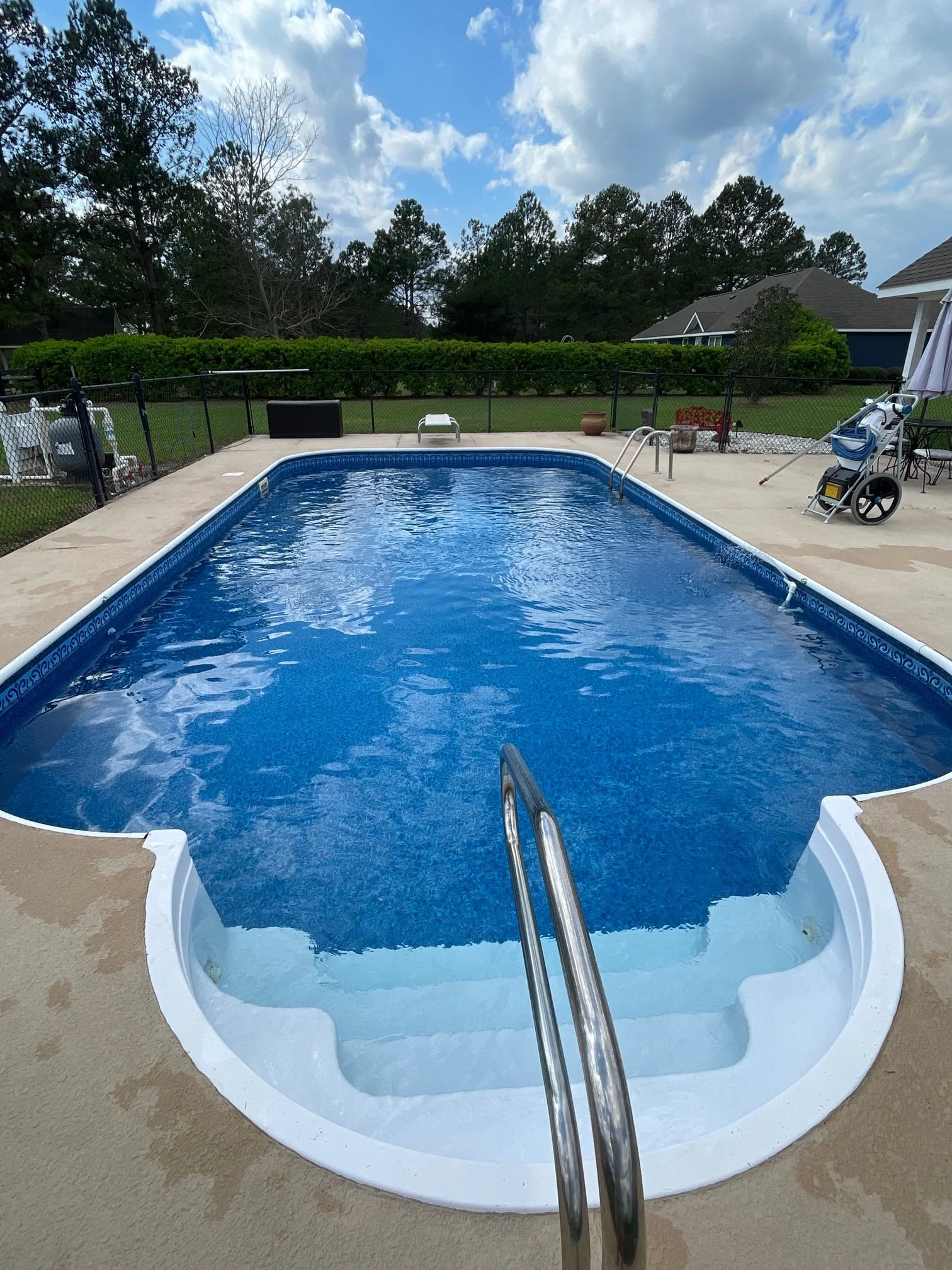 A backyard swimming pool with a metal ladder, surrounded by a concrete deck, with a lawn, trees, and houses in the background on a partly cloudy day.
