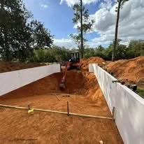 Construction site with white wall barriers and red dirt, excavation equipment in the background, and trees under a partly cloudy sky.