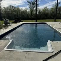 A rectangular in-ground swimming pool with a white pool deck in a backyard, surrounded by trees and a clear blue sky.