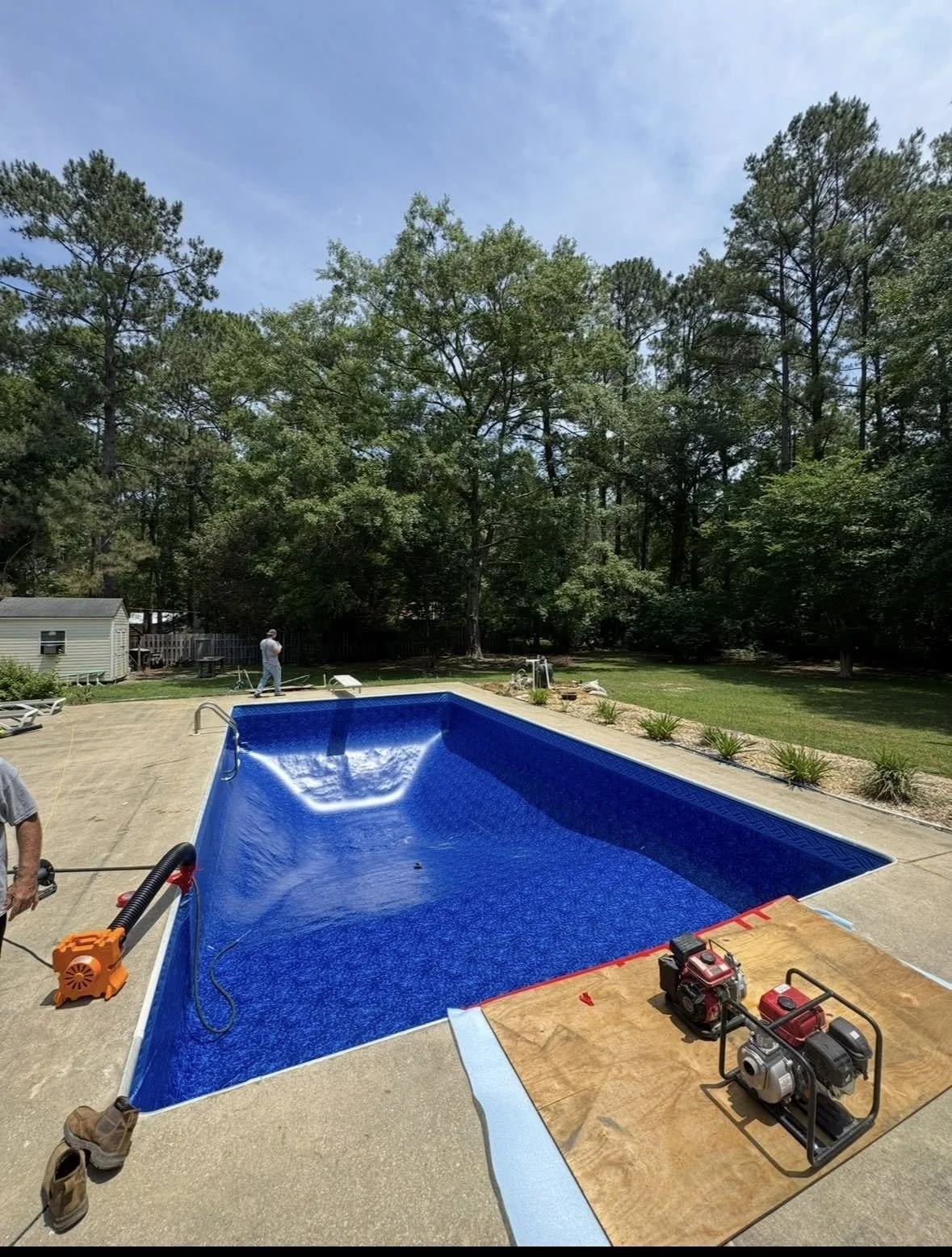 A backyard with a blue in-ground swimming pool, surrounded by a patio, with two men working on installation or maintenance. The background has tall green trees and a grassy lawn under a partly cloudy sky.