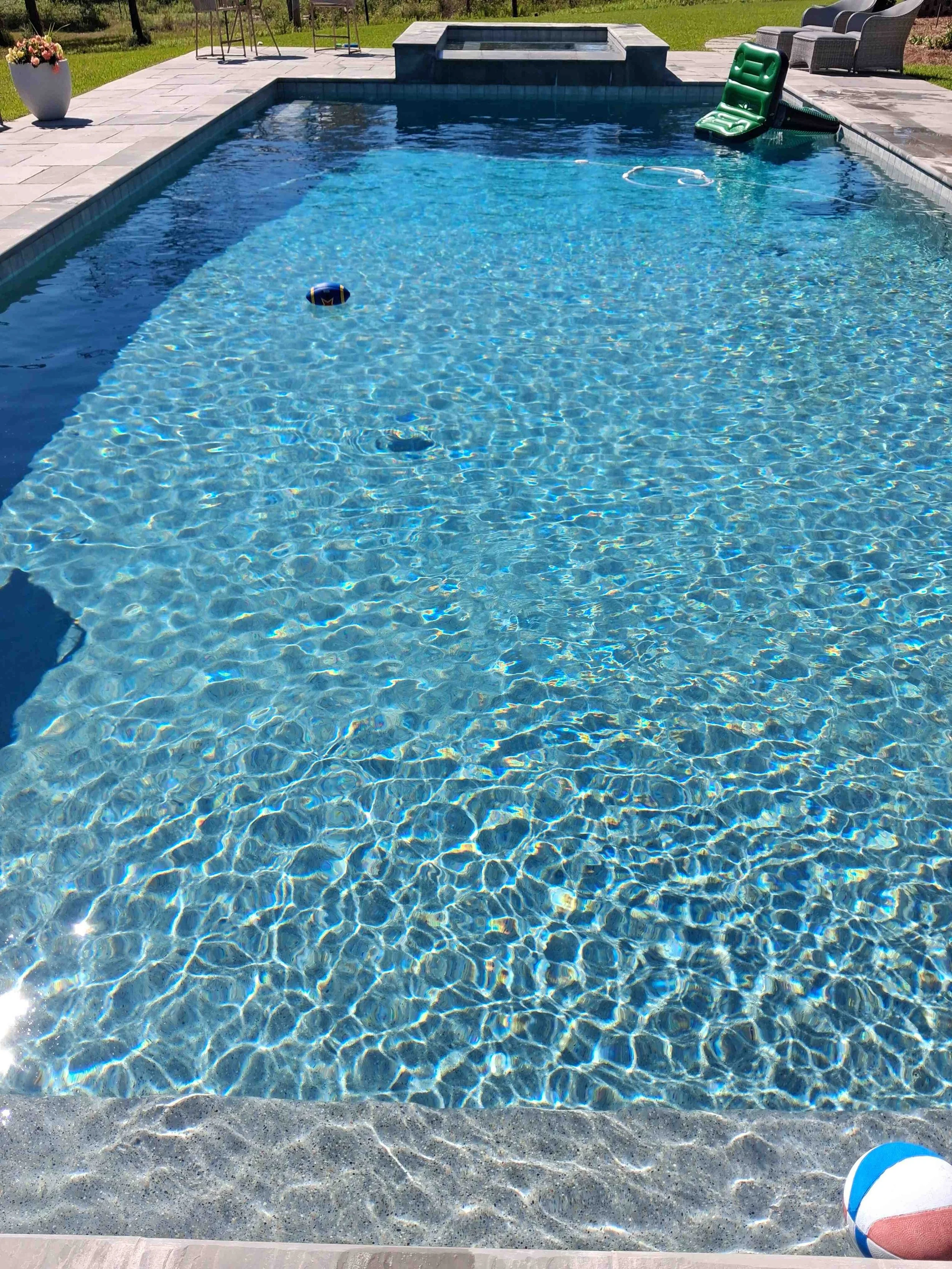 A backyard swimming pool with a partially submerged green lounge chair, a colorful beach ball on the pool deck, and outdoor furniture including chairs and a table in the background. The water in the pool is clear and blue, reflecting sunlight.