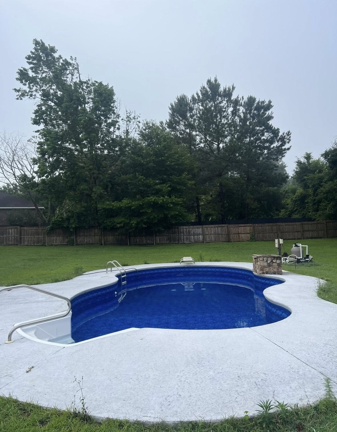 Backyard with a kidney-shaped swimming pool with a blue interior, surrounded by a white concrete deck. There is a pool ladder and a stone fire pit at the edge of the pool. In the background, there is a wooden fence, green grass, and tall trees, with 