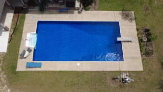 A backyard with a rectangular swimming pool surrounded by a concrete patio, with lawn and trees nearby.