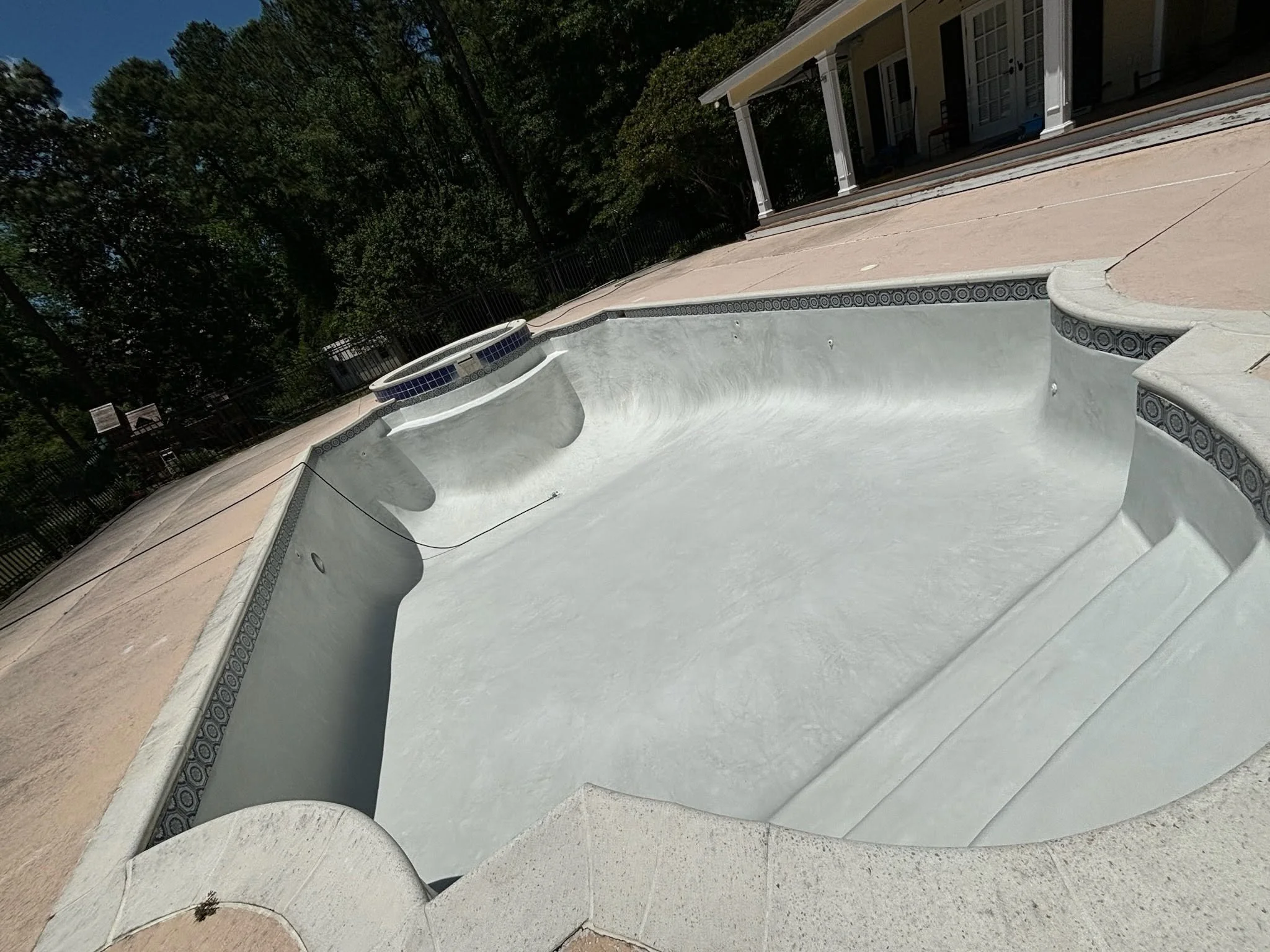 Empty swimming pool in backyard with house and trees in background.