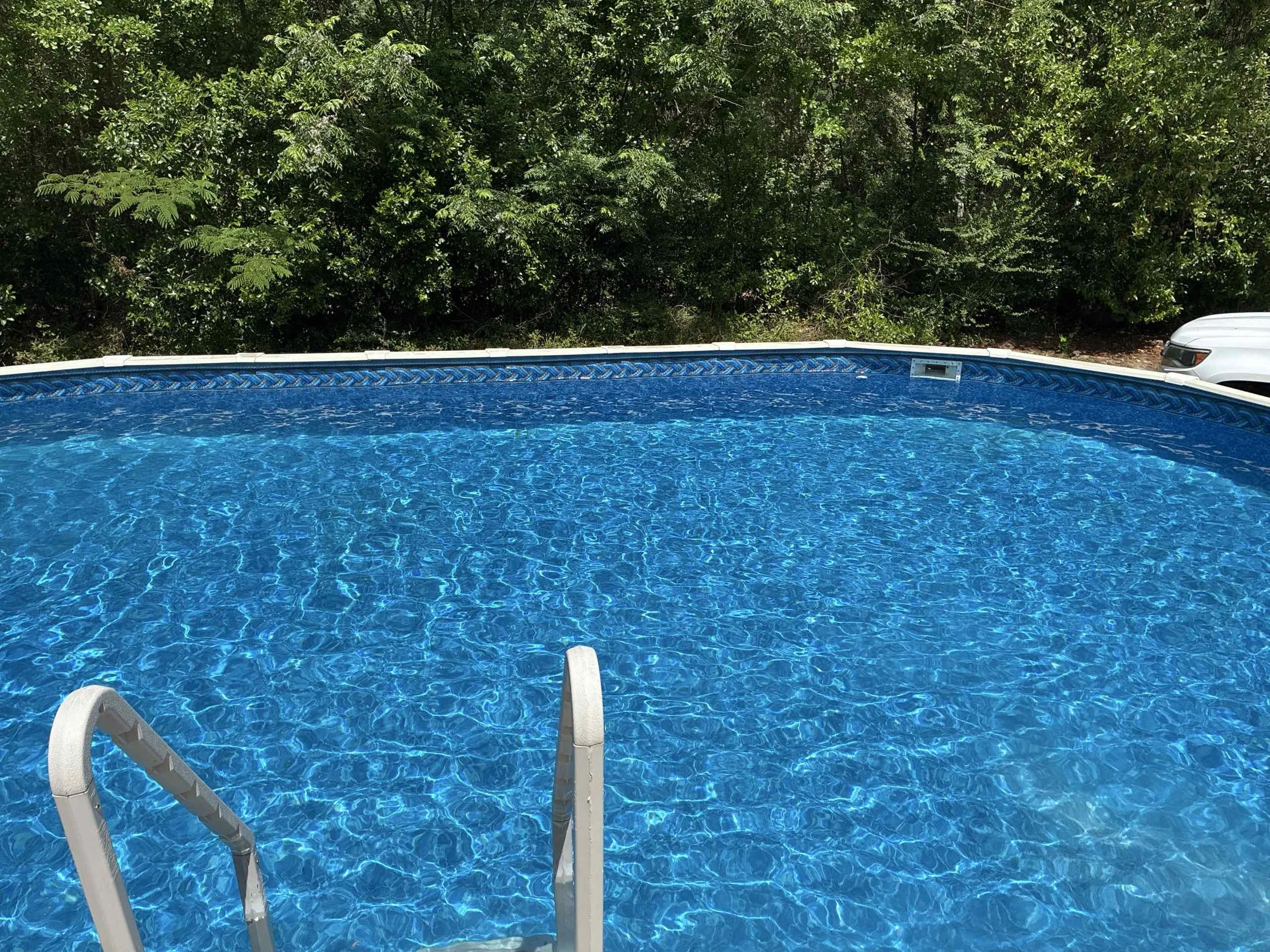 A swimming pool with blue water and white pool steps, surrounded by greenery and a white vehicle in the background.