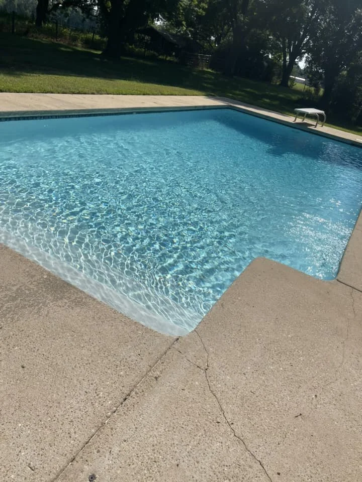 Deep end of a rectangular swimming pool with rippling water, surrounded by a concrete deck and a grassy backyard with trees.