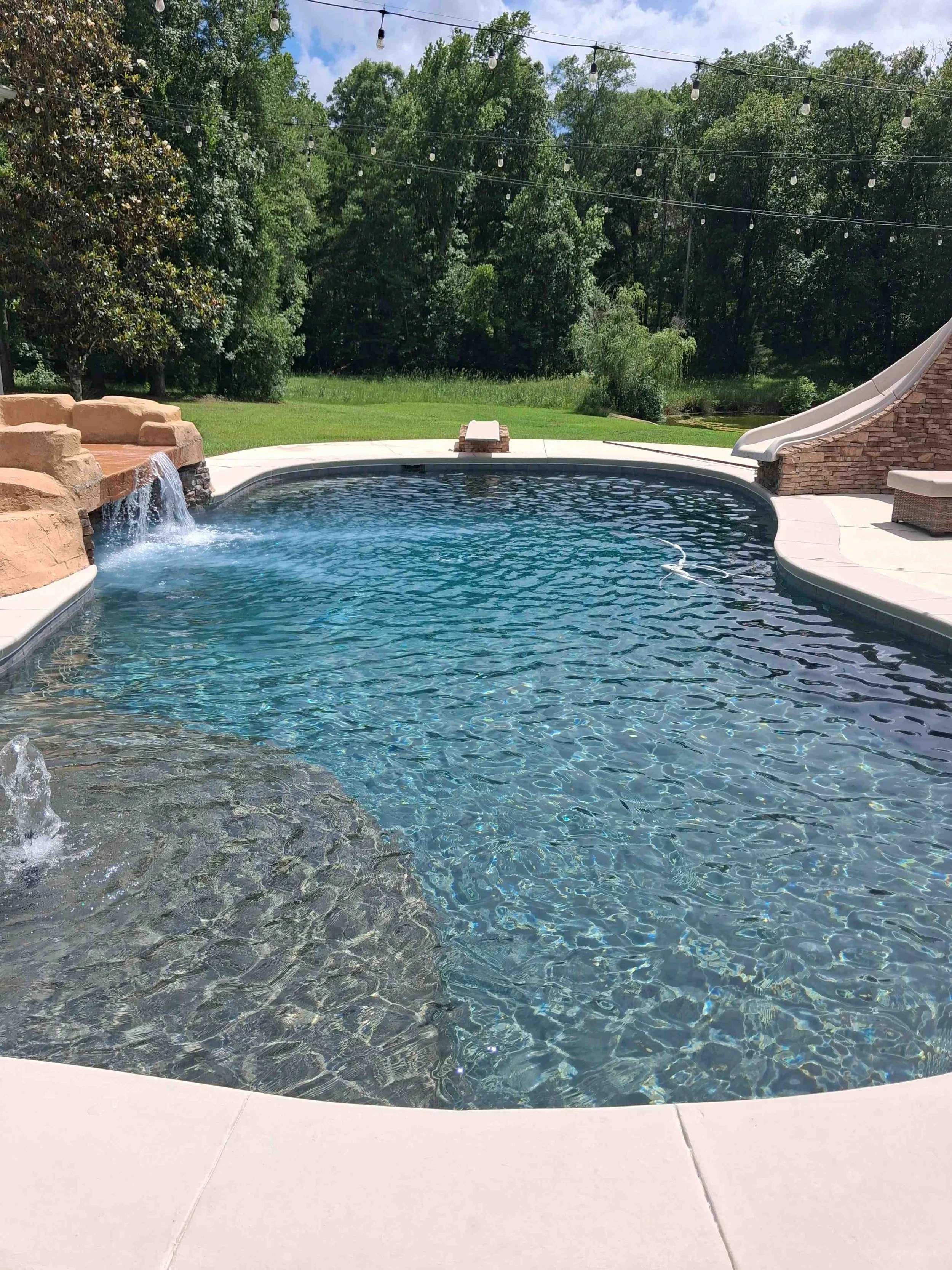A backyard swimming pool with a small waterfall feature on the left, surrounded by a white concrete deck. In the background, there are green lawns, trees, and string lights hanging overhead. A slide is visible on the right side.