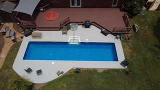 A backyard pool with water, surrounded by a concrete deck, outdoor furniture, and an adjacent deck with a patio umbrella.