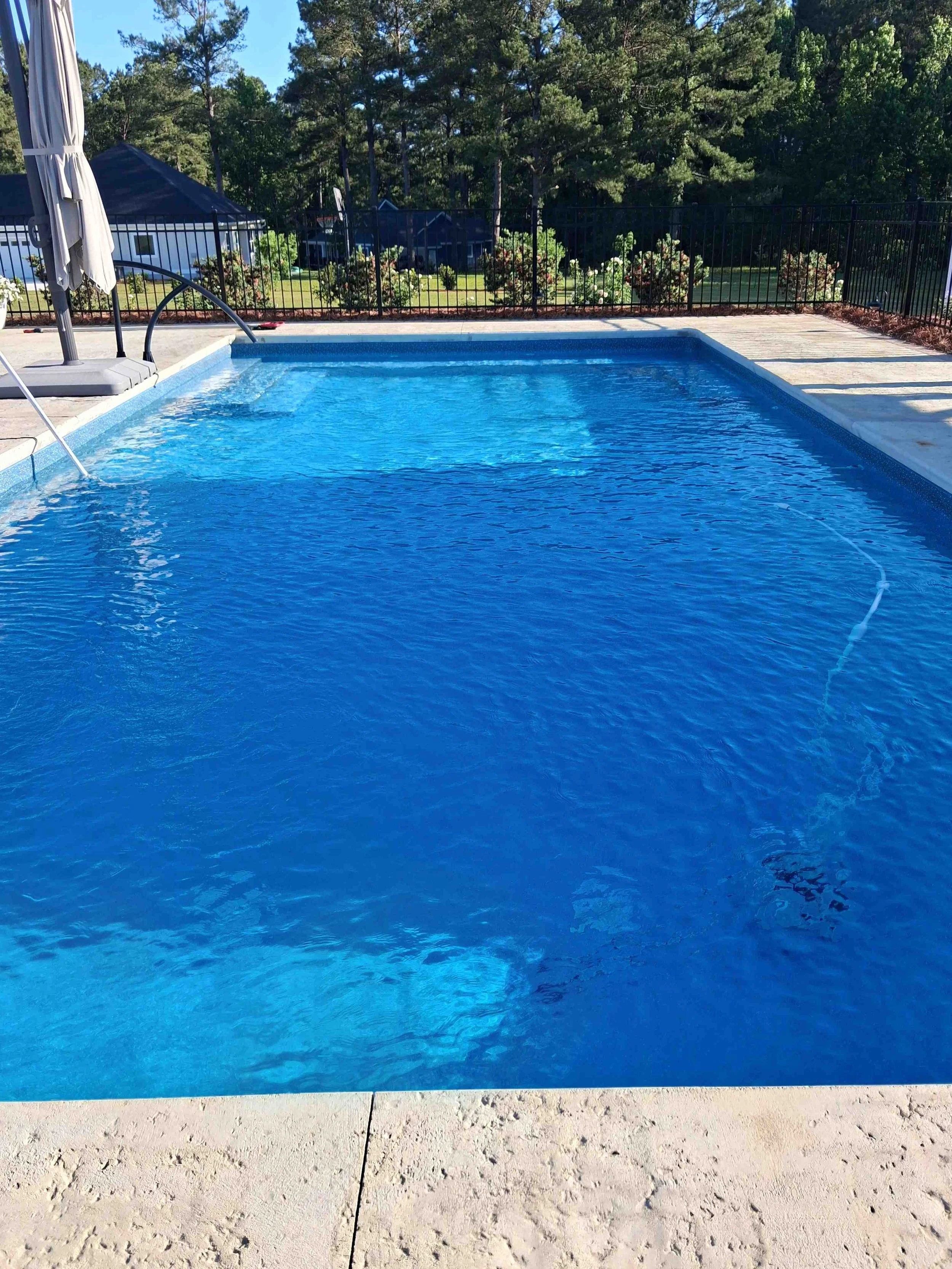 A backyard swimming pool with blue water, surrounded by a concrete deck and black metal fence, overlooking a green lawn with flowering bushes and trees.