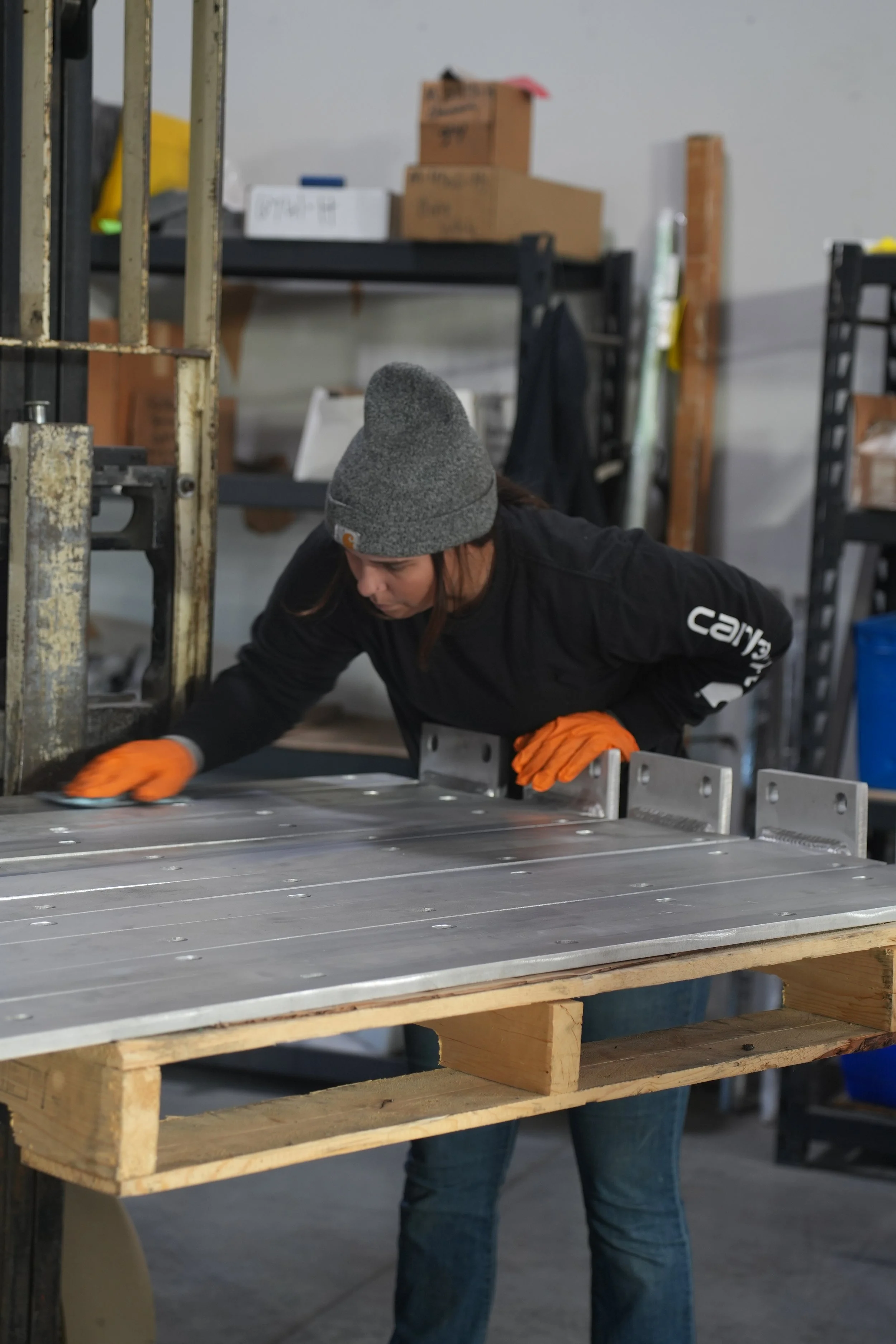 A woman wearing a gray beanie, black sweatshirt, and orange gloves is working with metal sheets at a workbench in a workshop. Shelves with boxes and tools are in the background.