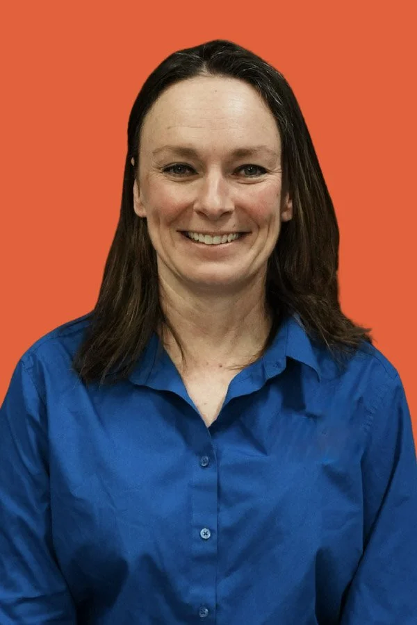 A woman with shoulder-length dark hair, smiling, wearing a blue button-up shirt, against a solid orange background.