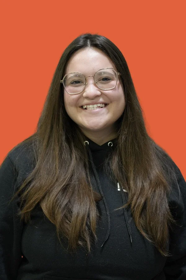 A young woman with long brown hair, glasses, and a black hoodie smiling, standing in front of an orange background.