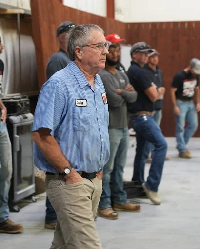A group of men standing in line indoors, with one man in the foreground wearing glasses, a blue plaid shirt with a name tag that says 'Craig,' and beige pants, appearing thoughtful.