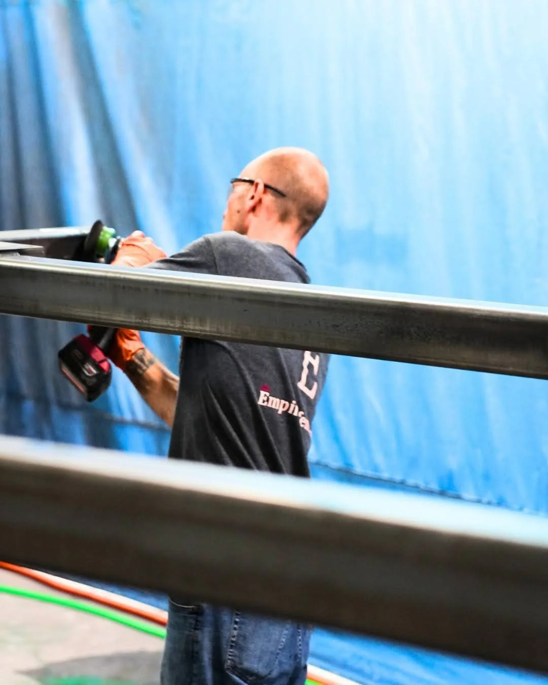 A man with glasses and a shaved head, wearing a dark shirt, is holding a cordless drill and working with a large metal pipe against a blue backdrop.