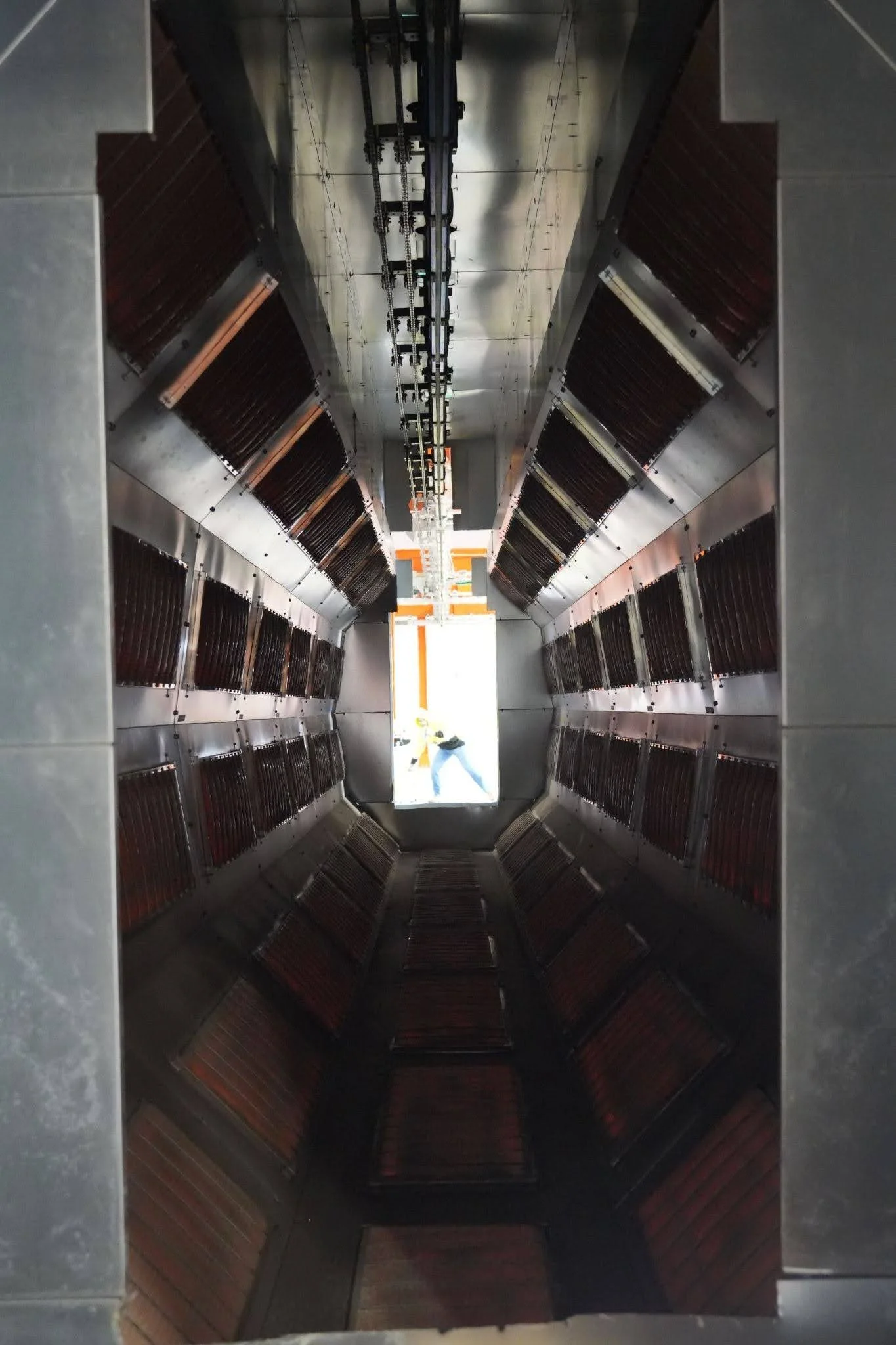 The interior of a stainless steel chimney with descending openings containing black slats or panels, and a person visible at the far end near an orange structure.