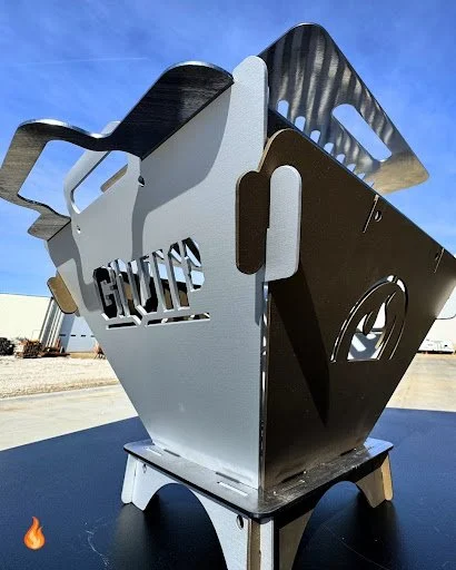 A large, modern metal outdoor fire pit with a unique cutout design and a decorative handle, situated outdoors on a sunny day.
