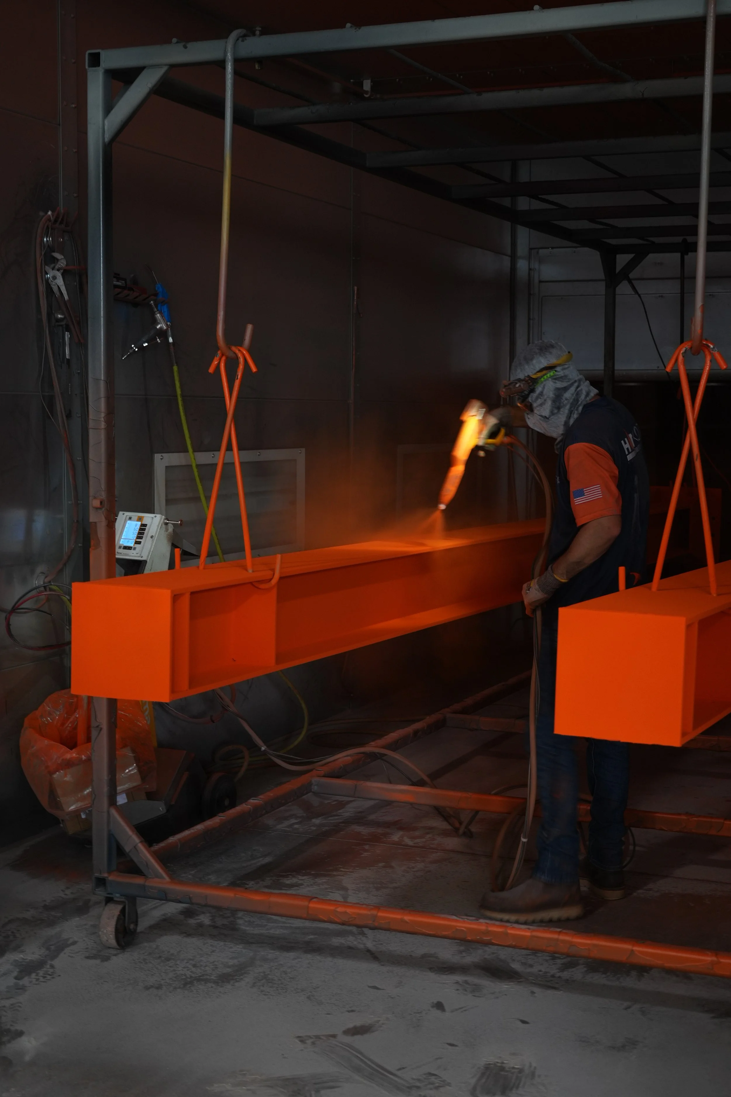Worker in protective gear welding orange metal structures inside an industrial workshop with tools hanging on the wall. The worker has a flame coming out of a tool as if welding.