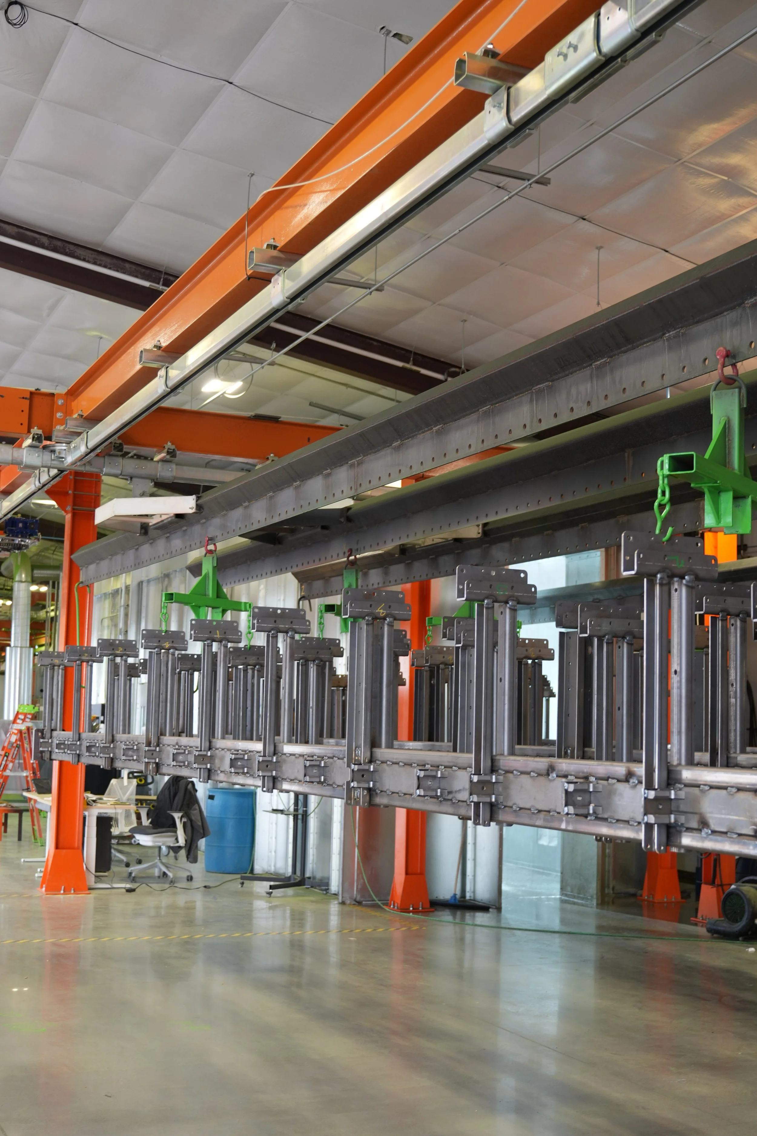 Indoor industrial workspace with large orange and gray metal machinery and equipment, including metal rails and green clamps, in a factory or manufacturing environment.