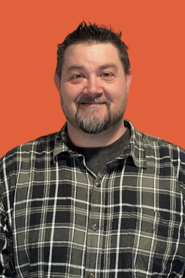 A man with short dark hair and a beard, wearing a black and green plaid shirt, stands in front of a solid orange background.