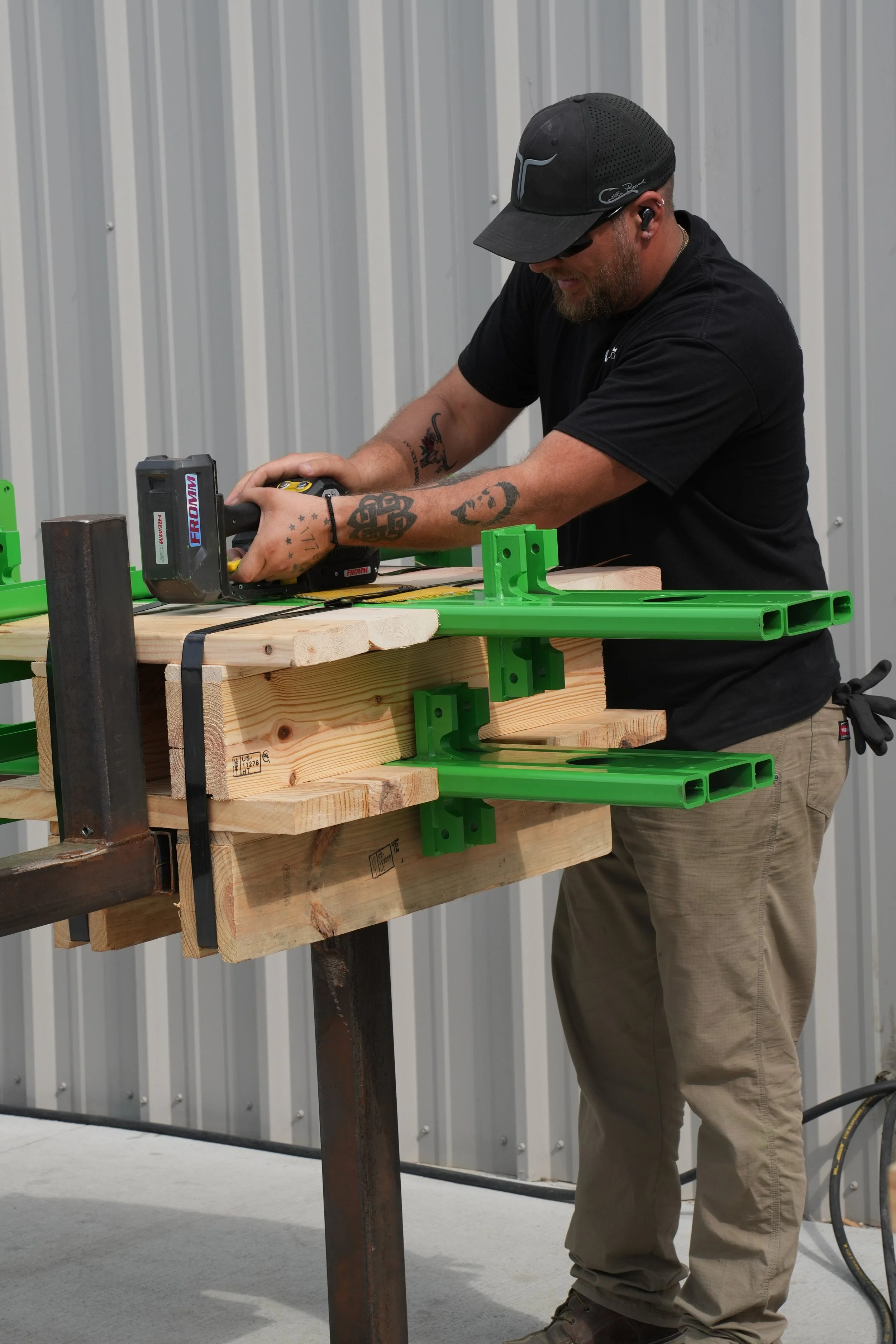 A man working on a woodworking project with a cordless nail gun, wearing safety glasses and a black cap, in a workshop with metal siding in the background.