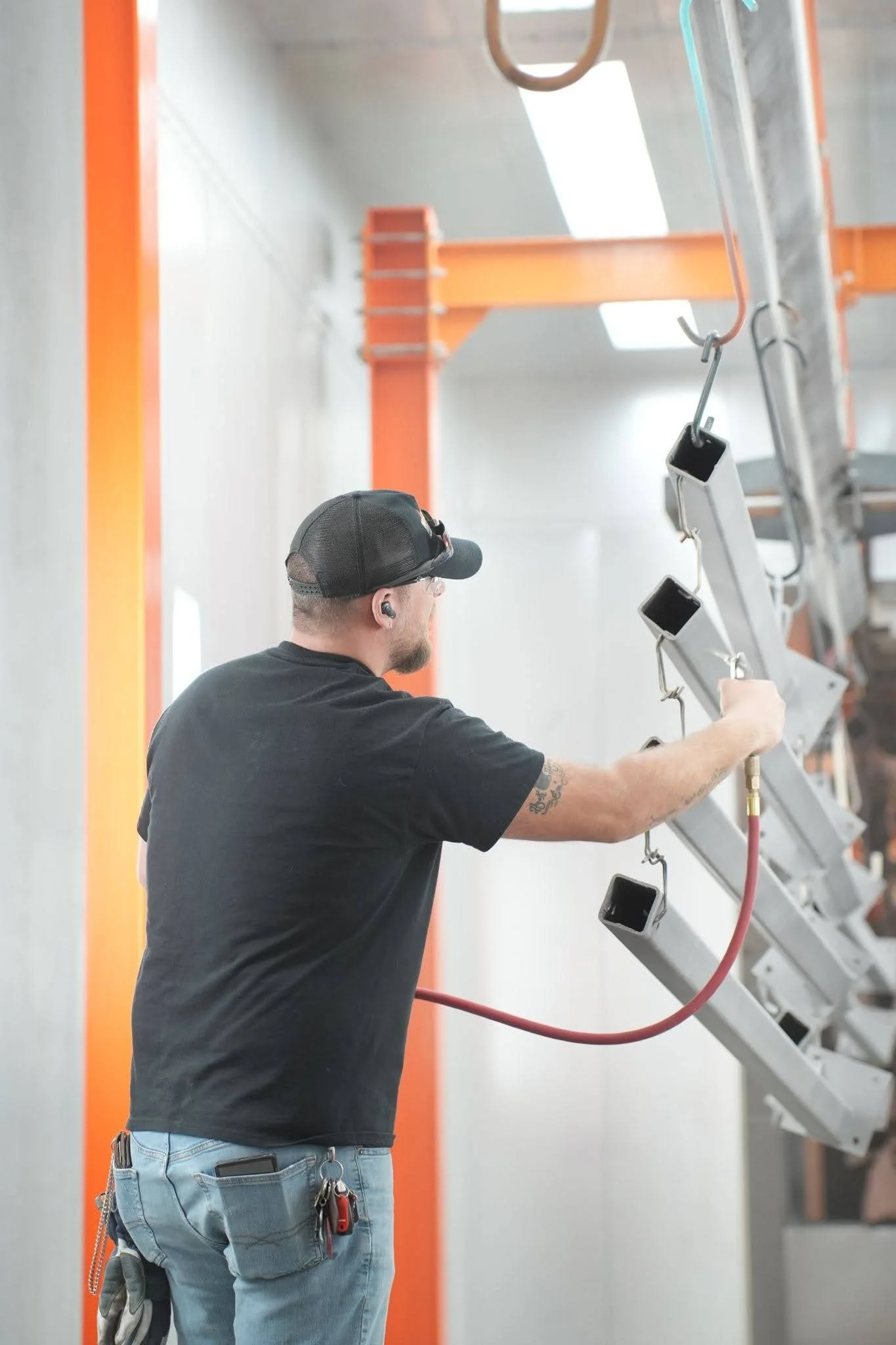 A man wearing a black cap, glasses, and ear protection working on a ladder in an industrial setting with orange metal beams and a gray wall.