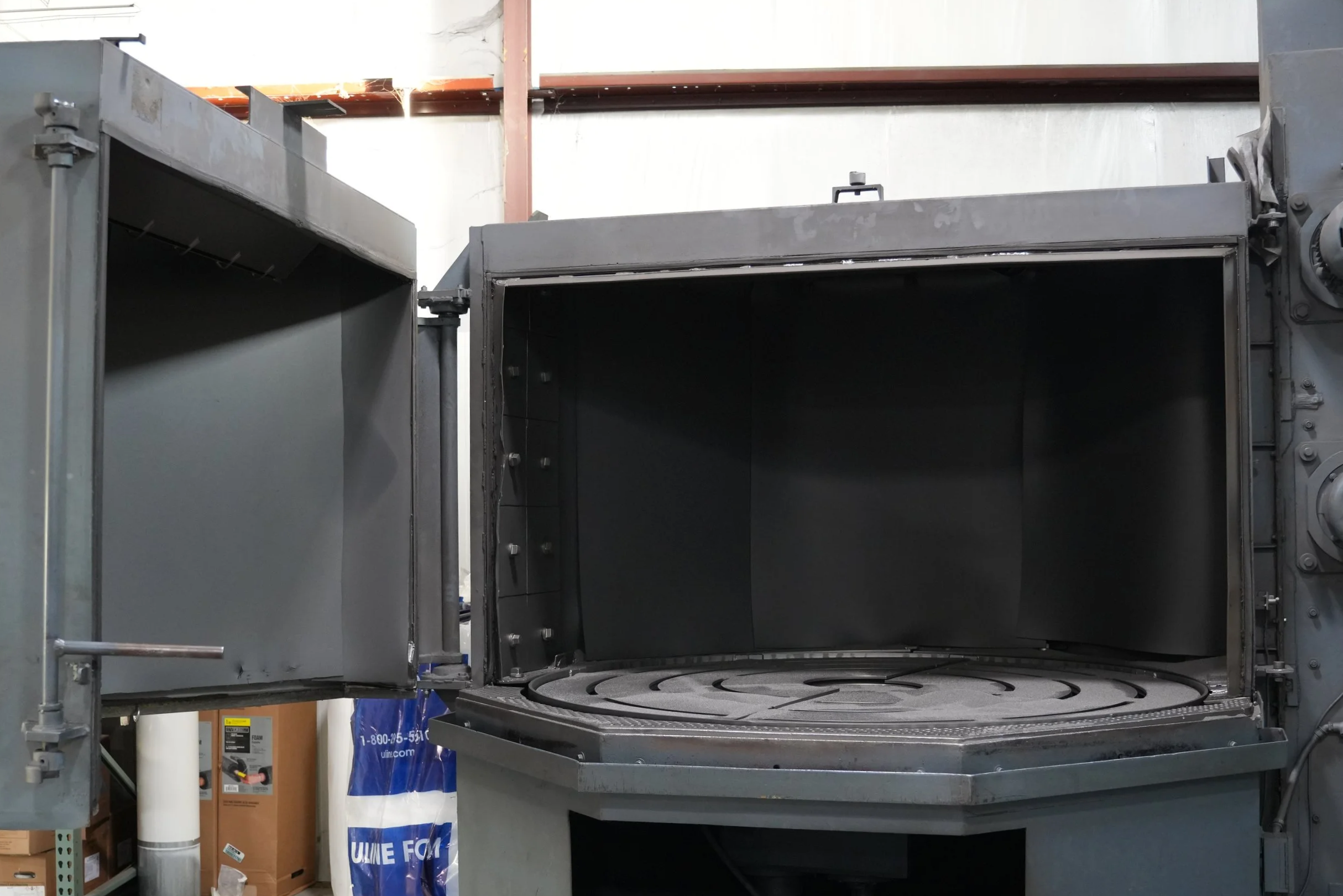 Empty industrial oven with open doors, metal interior, and a circular tray inside, situated in a warehouse or workshop setting with pipes, boxes, and equipment visible around.