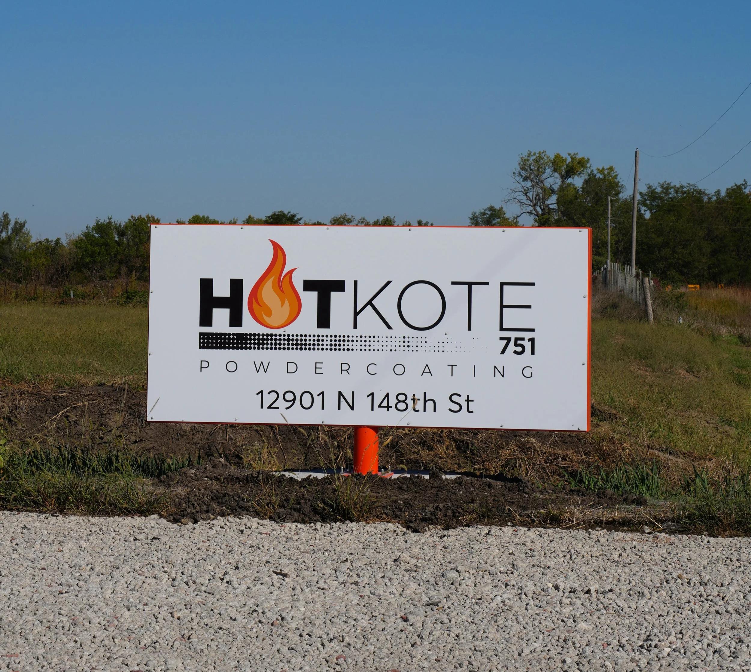 Sign with a fire flame graphic and the words 'HOTKOTE POWDERCOATING' along with an address and phone number, placed outdoors on a gravel surface with trees and utility poles in the background.