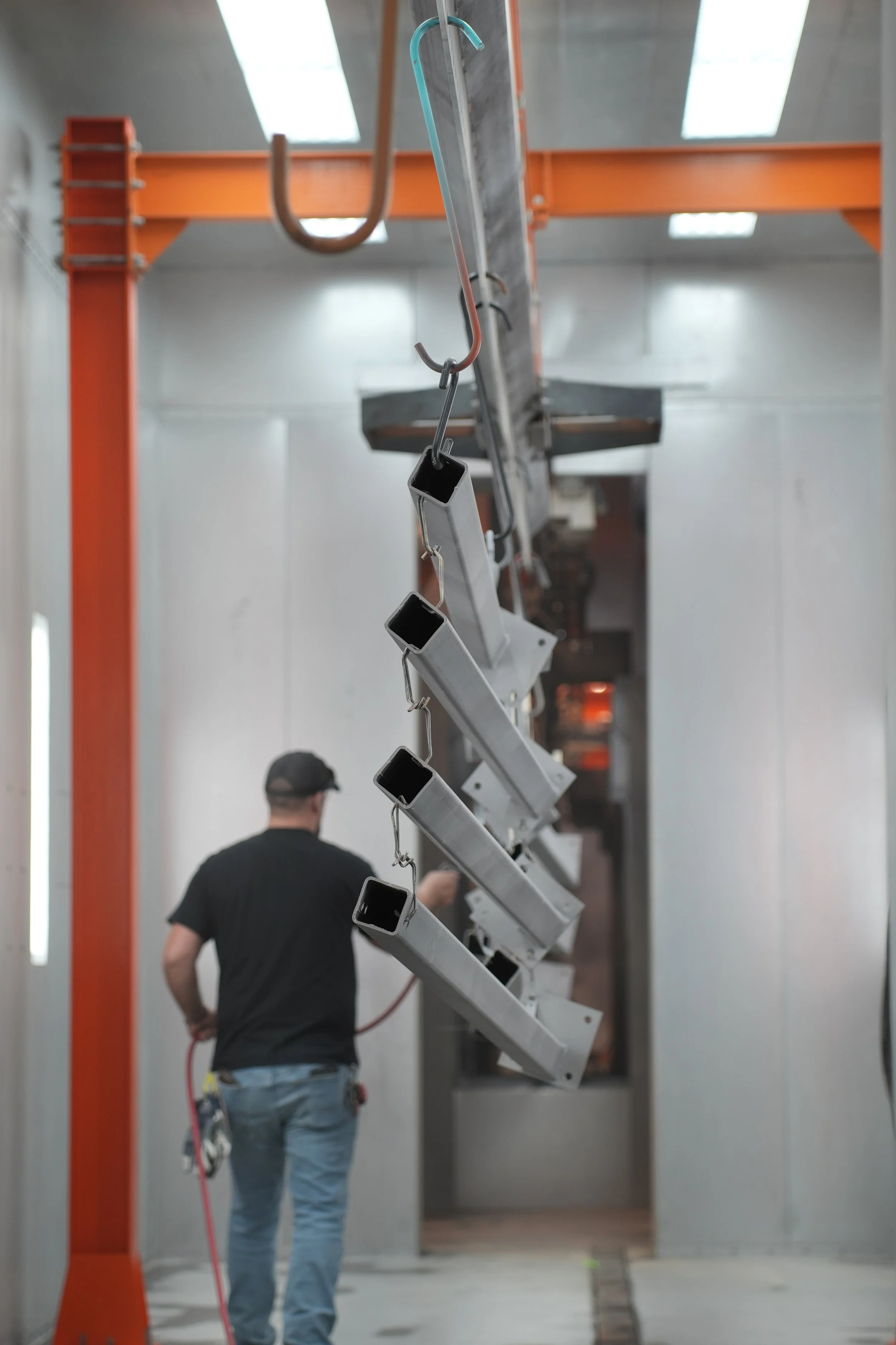 A man in a black T-shirt and jeans is working on installing or repairing a large lighting fixture in an industrial or commercial space. The ceiling has large skylights, and a prominent orange metal beam supports the ceiling structure.
