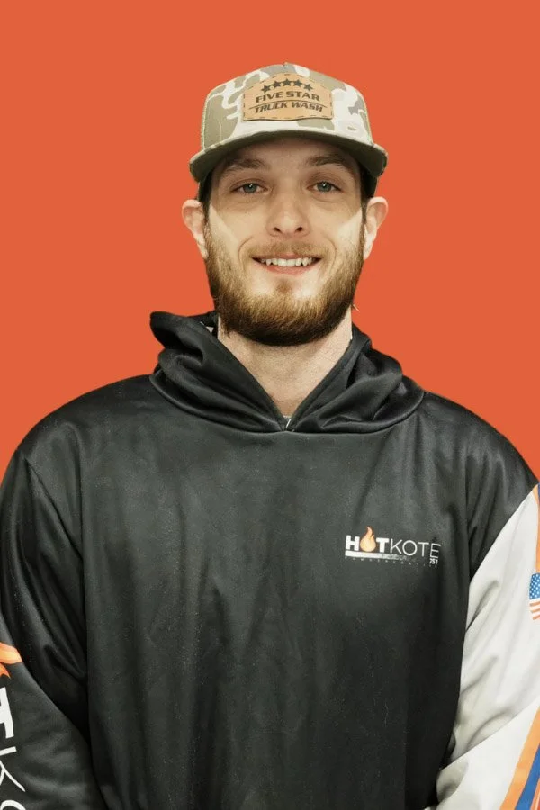 Young man with a beard smiling, wearing a black hoodie with a logo that says "HOKOTE" and an American flag patch on the sleeve, and a camouflage cap with a patch that reads "Five Star Truck Wash," standing against an orange background.