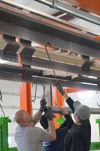 Two men working on installing or repairing a large metal structure, using tools under a ceiling with orange beams and green structural supports.