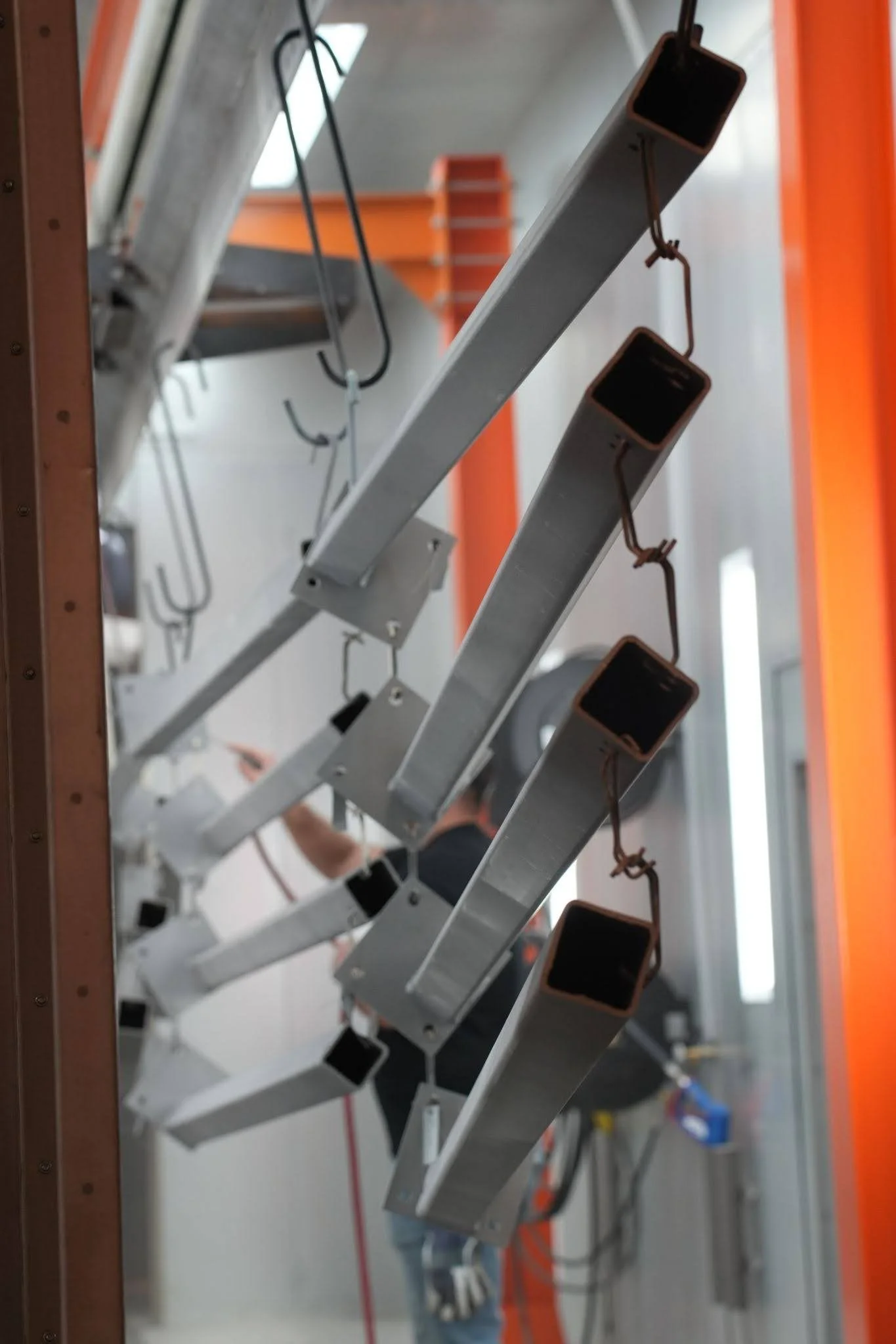 Close-up view of a metal structure with clipped wires or cables, possibly in an industrial or construction setting, with blurry background including orange and white walls and equipment.