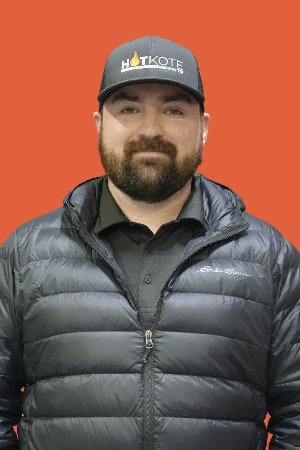 A man with a beard and mustache wearing a black cap with a logo, a black collared shirt, and a dark gray puffer jacket, standing against an orange background.
