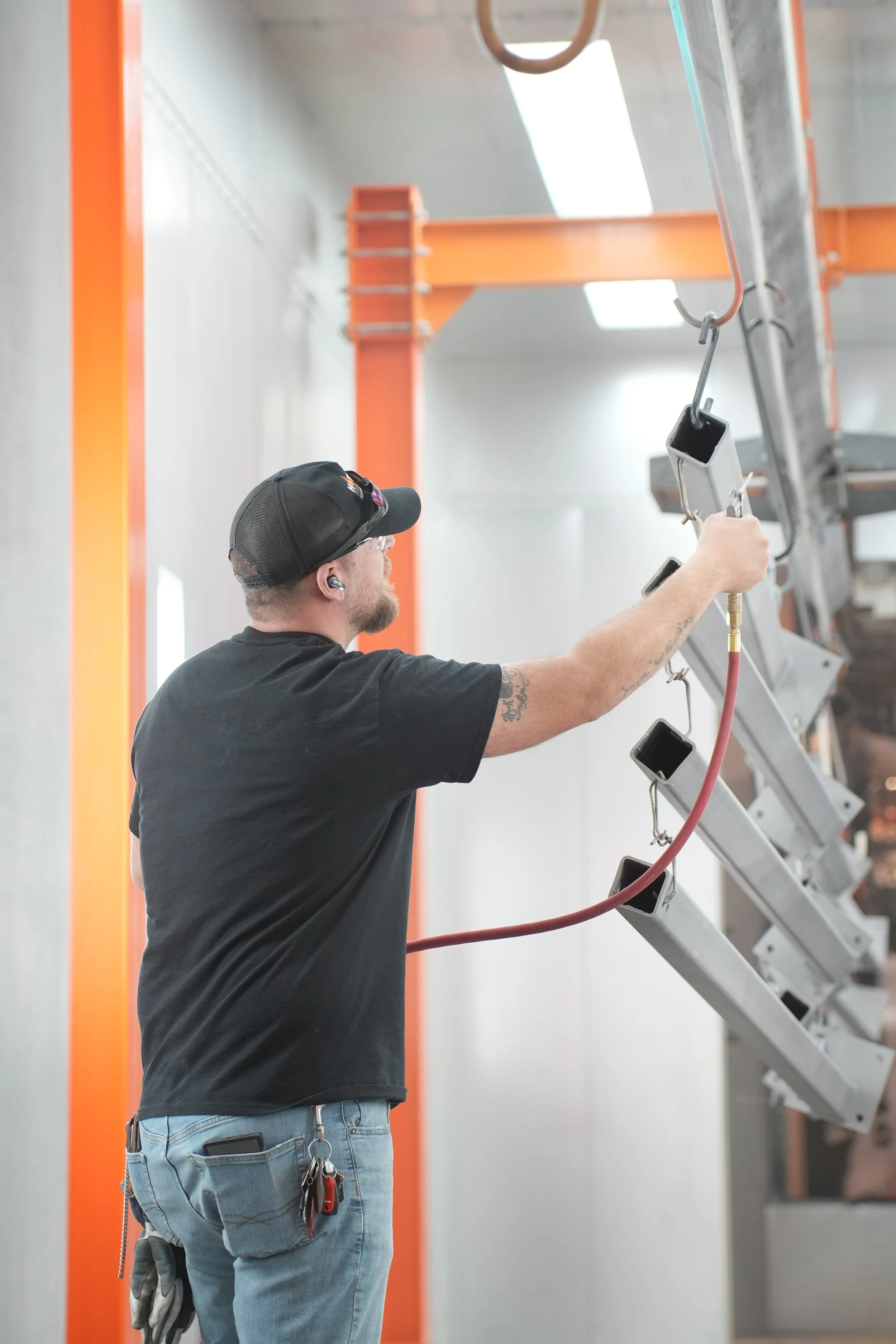 A man wearing a black cap, glasses, and earbuds is reaching up to adjust or install equipment on a metal ladder in an industrial or warehouse setting. The background includes orange beams and a gray wall.