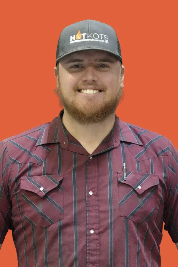 A young man with a beard smiling, wearing a gray hat with a "HIT KOTE" logo, and a maroon striped button-up shirt, standing against an orange background.
