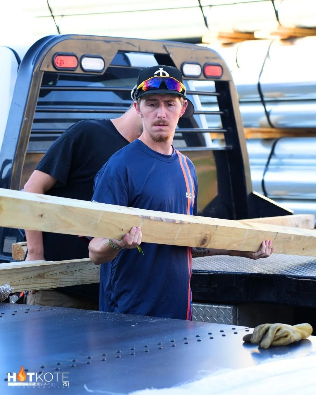 A young man wearing a black cap with a white symbol, colorful sunglasses on his cap, and a blue sports shirt, holding a piece of wood while standing near a truck with metal and wood supplies.