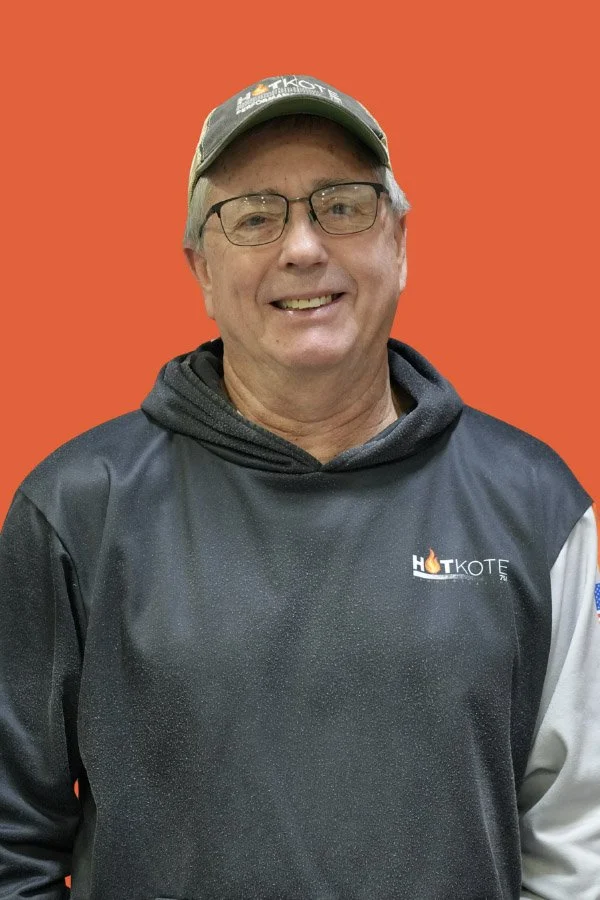 A smiling middle-aged man wearing glasses, a green cap, and a black sweatshirt with the 'HOTKOTE' logo, standing against an orange background.