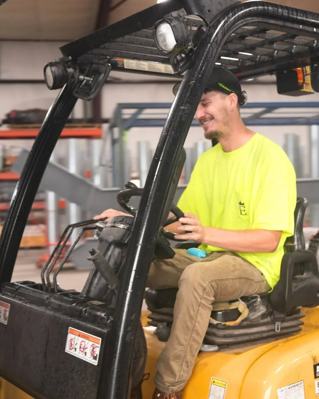 Young man in a bright yellow safety shirt and khaki pants driving a yellow forklift inside a warehouse, smiling while looking at the controls.