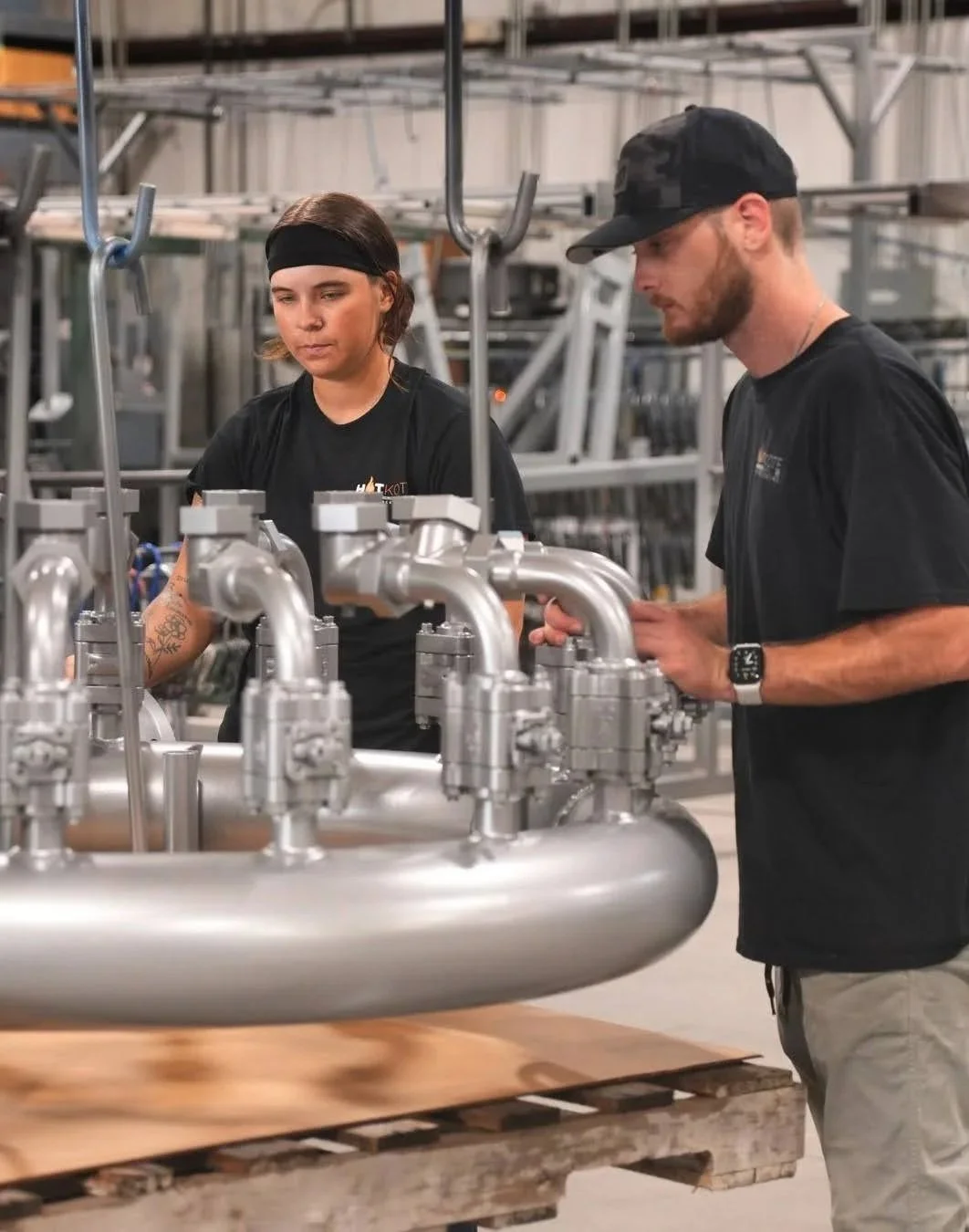 Two workers, a woman with a headband and a man wearing a cap, inspecting a stainless steel industrial pipe system in a manufacturing or warehouse setting.