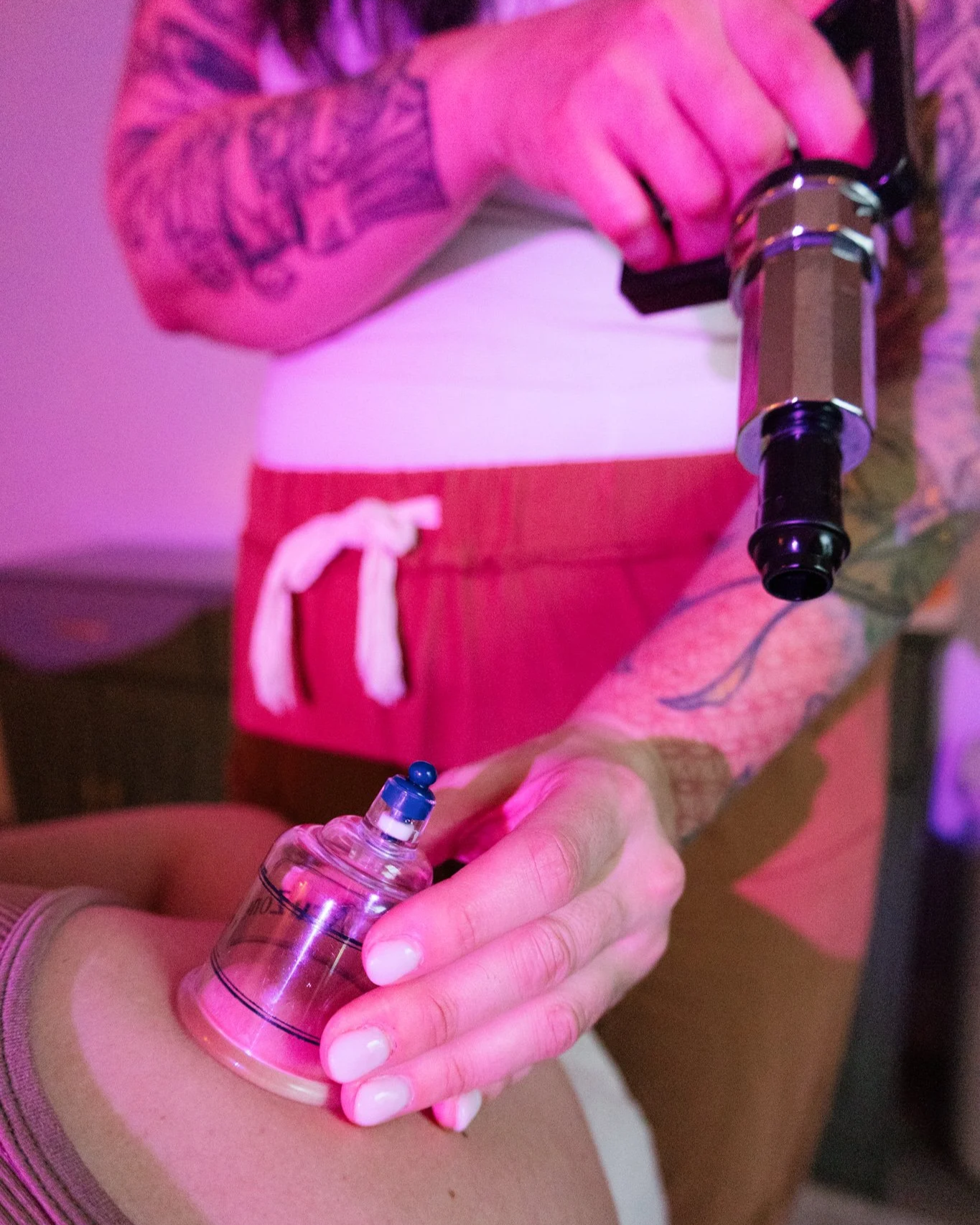 Person holding a syringe and a small vial, preparing for an injection. The person is wearing rings and has tattoos on their arm, with a pink and purple lighting ambiance.