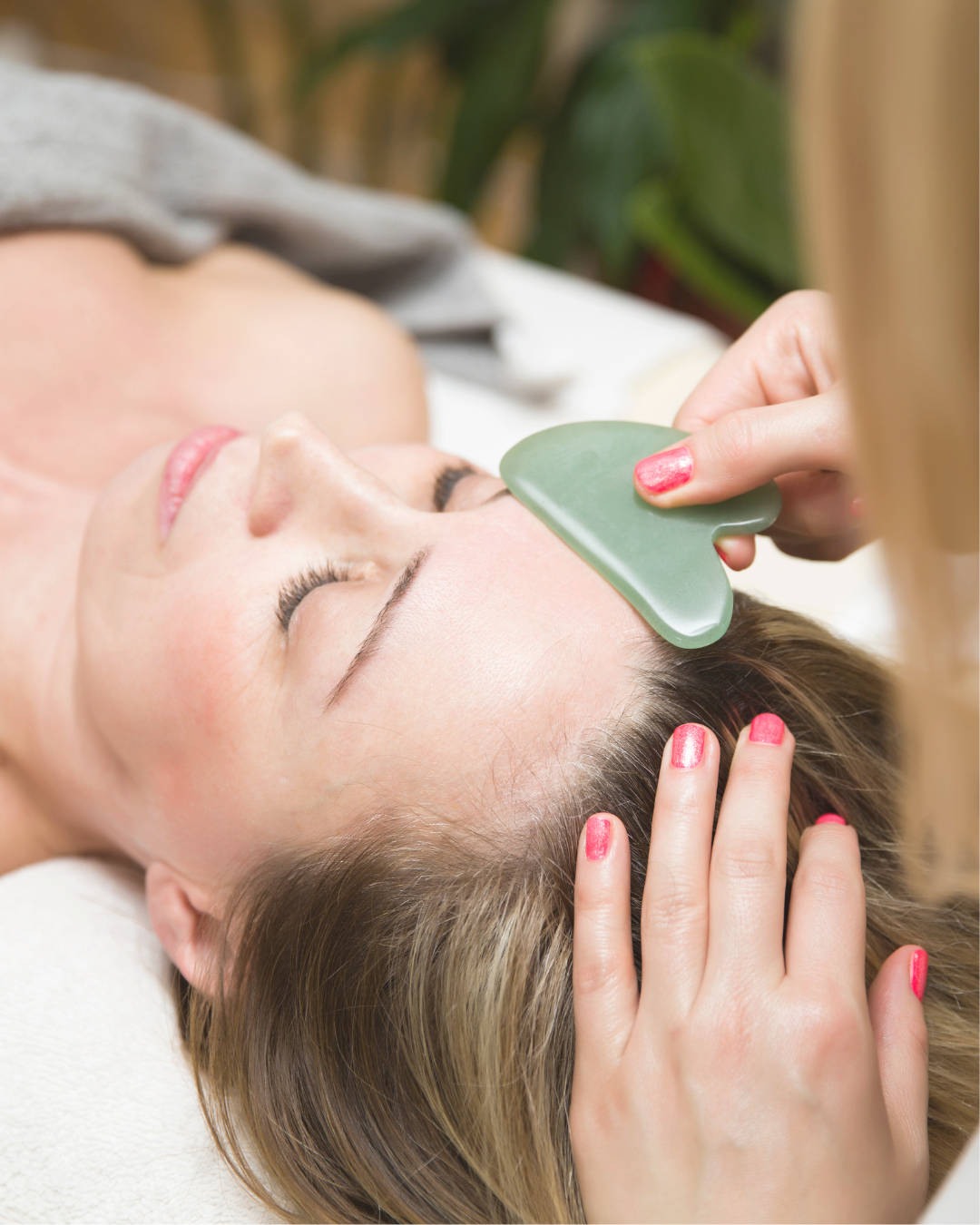 A woman receiving a facial treatment with a jade roller on her forehead, lying down with her eyes closed.