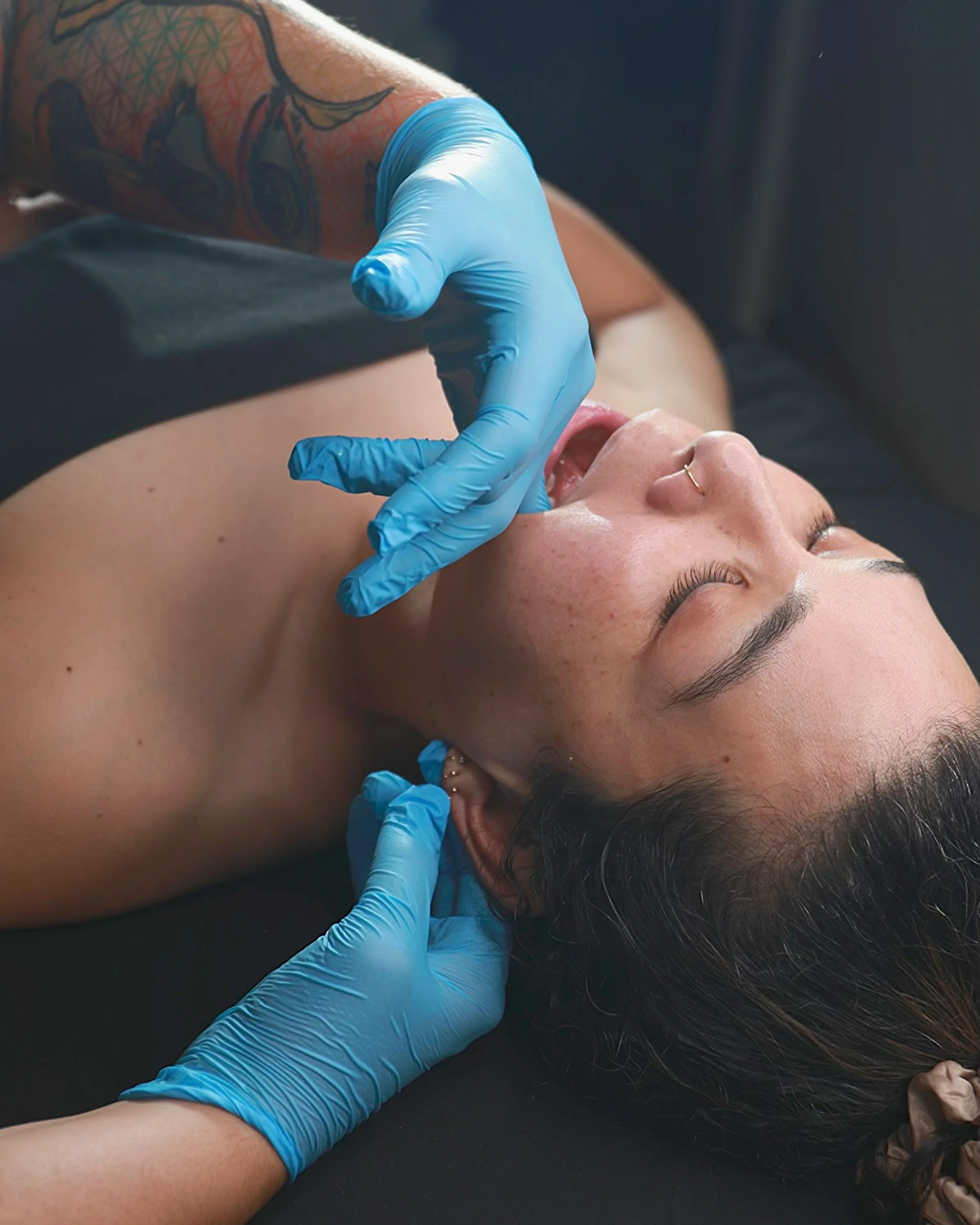 Close-up of a person lying down with eyes closed, receiving a tattoo on their upper arm from a tattoo artist wearing blue gloves.