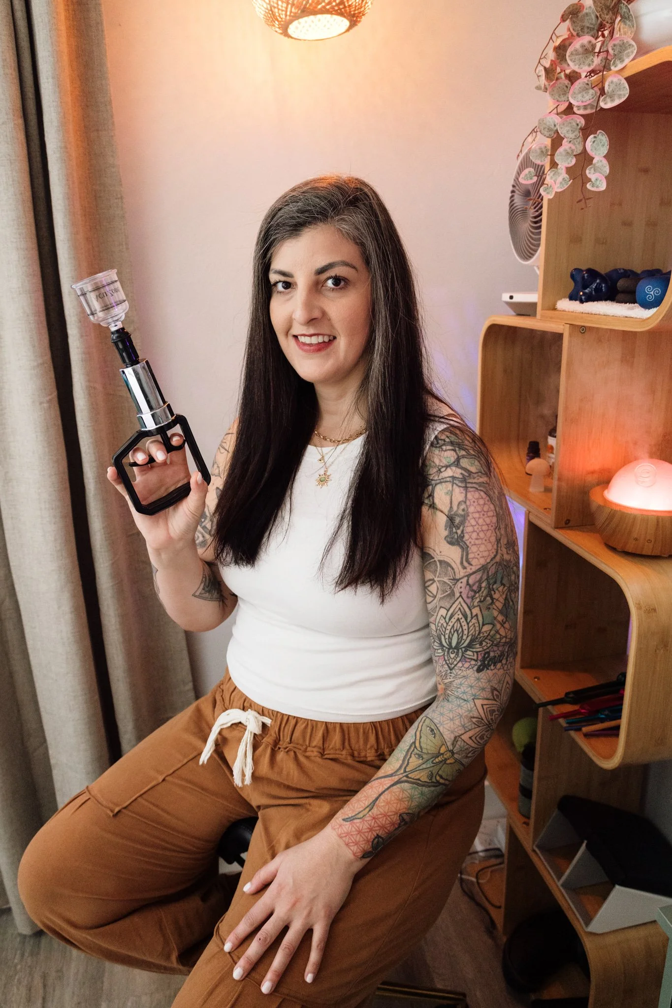 A woman with long dark hair and tattoos on her arms, wearing a white tank top and brown pants, is sitting on a chair and holding a glass piece of a vape mod or vaporizer. She is smiling and looking at the camera in a cozy room with wooden shelves, decorative items, and warm lighting.
