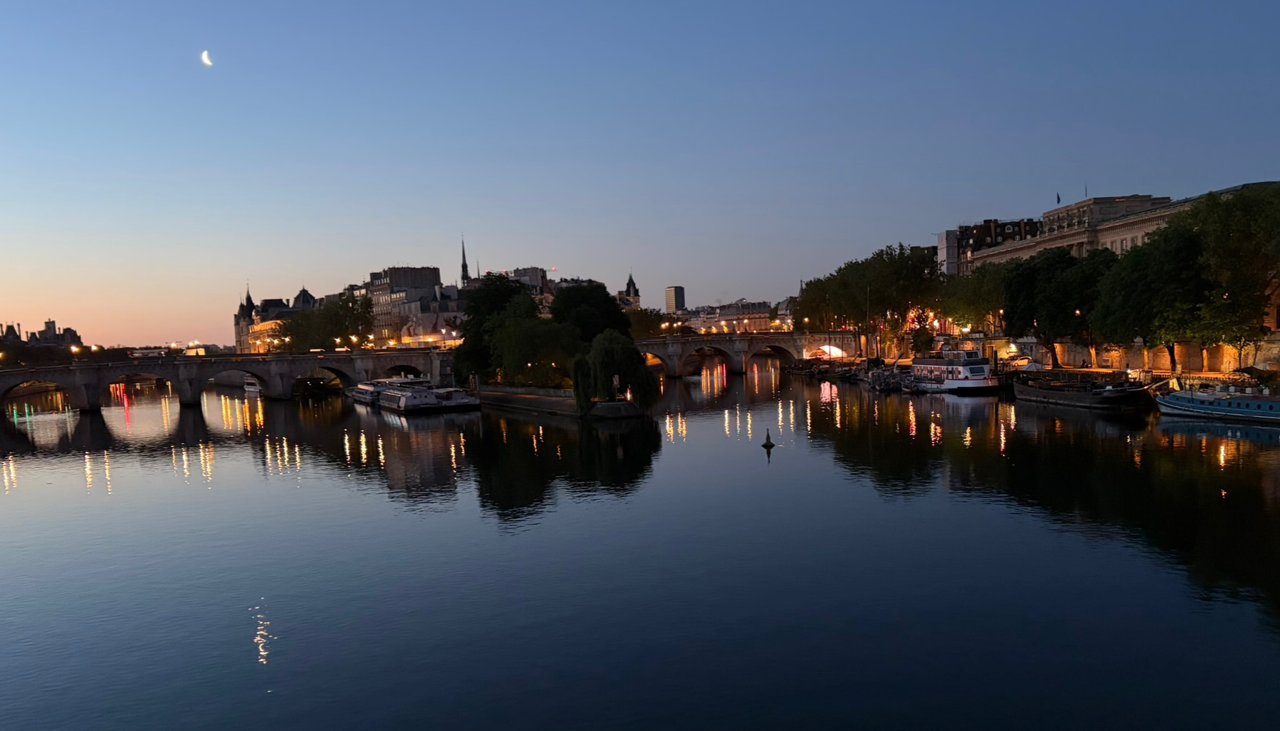 Nighttime view of a cityscape with a river in the foreground, boats docked along the riverbank, illuminated buildings lining the street, and a bridge over the river. The sky is clear with a crescent moon visible.