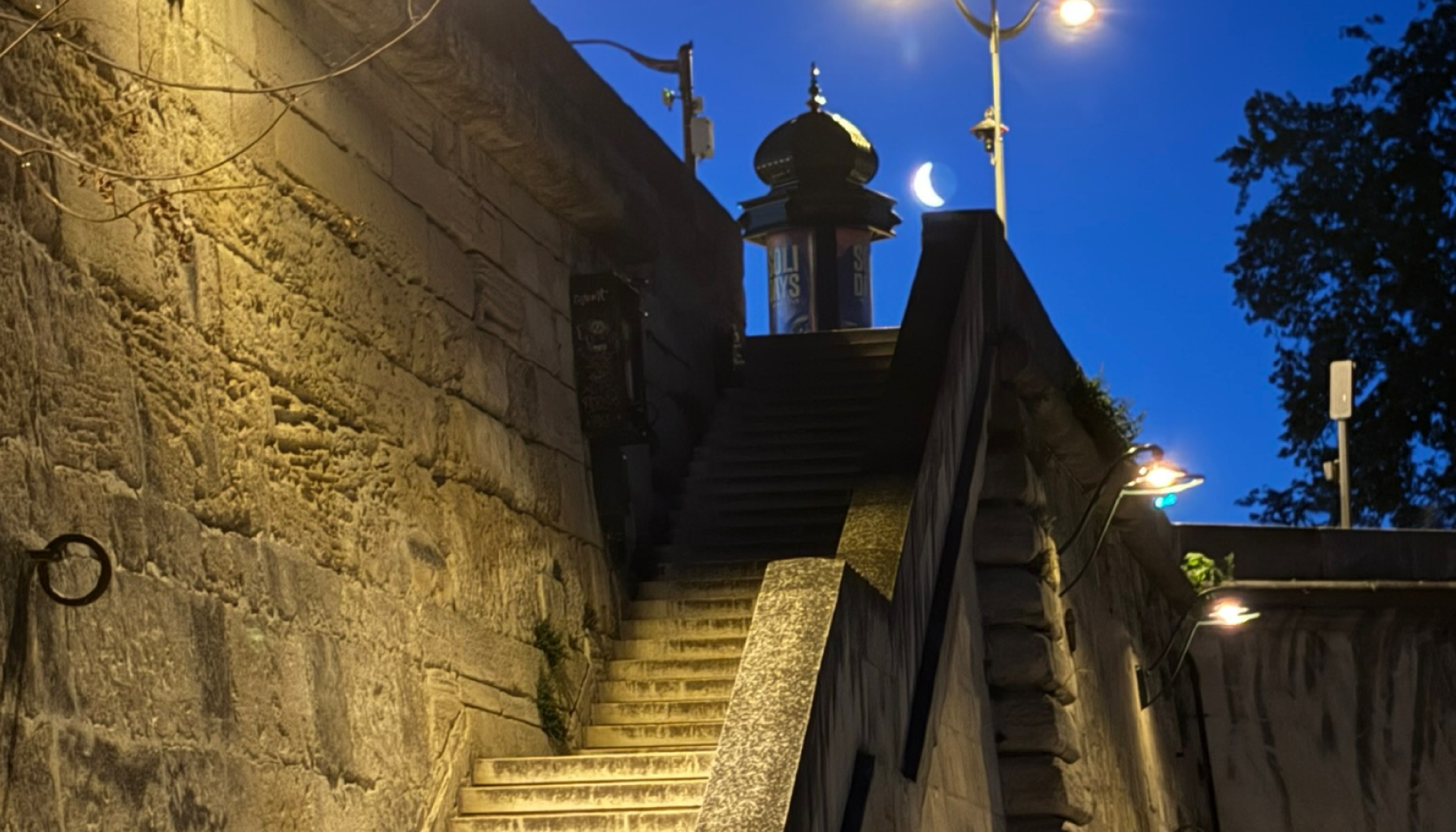 Stone staircase leading up next to a stone wall with outdoor lights and small plants, with a dark blue evening sky in the background, featuring a crescent moon and streetlights.